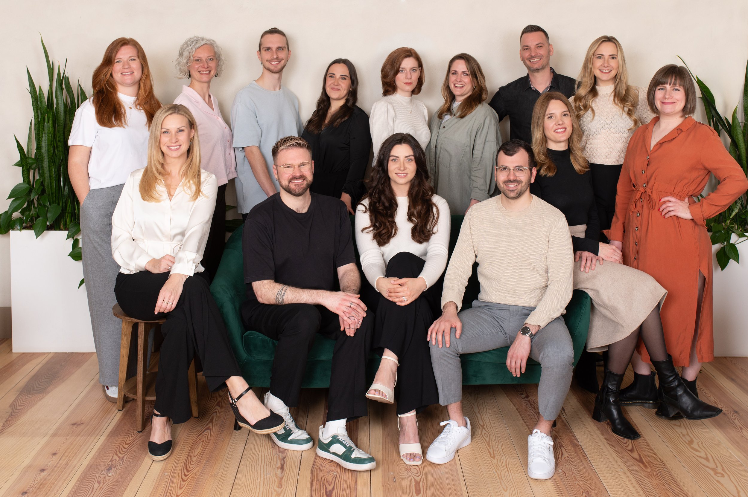 Gruppenfoto einer multikulturellen Arbeitsgruppe vor weißer Wand mit Pflanzen, 3 Personen sitzen auf einem grünen Sofa, 10 Personen stehen im Hintergrund.