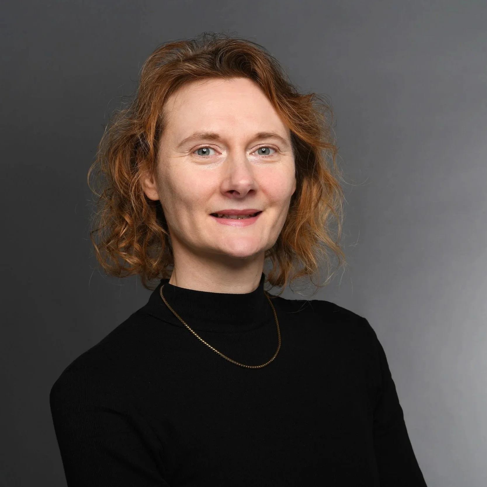 Porträt einer Frau mit roten, lockigen Haaren, schwarzem Rollkragenpullover und einer goldenen Kette vor grauem Hintergrund.
