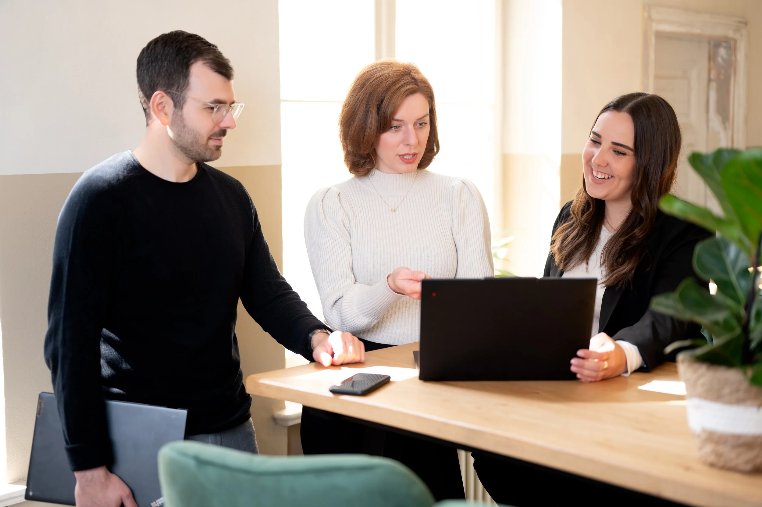 Drei Menschen diskutieren um einen Laptop in einem Büro, eine Frau zeigt auf den Bildschirm, alle lachen und wirken interessiert.