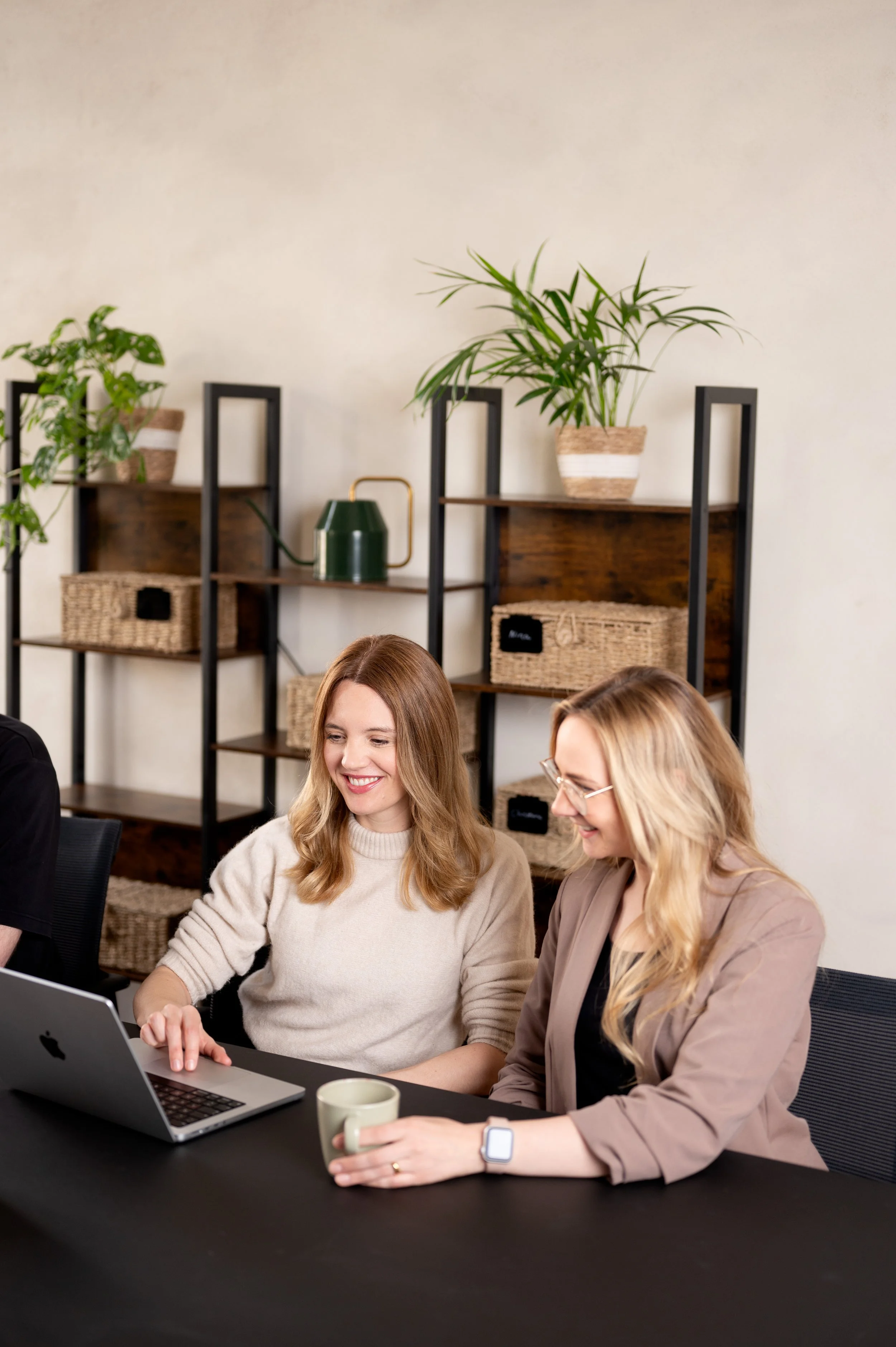 Zwei Frauen sitzen an einem schwarzen Tisch, schauen auf einen Laptop, lachen und haben eine Tasse in der Hand, im Hintergrund Pflanzen und dekorative Regale.