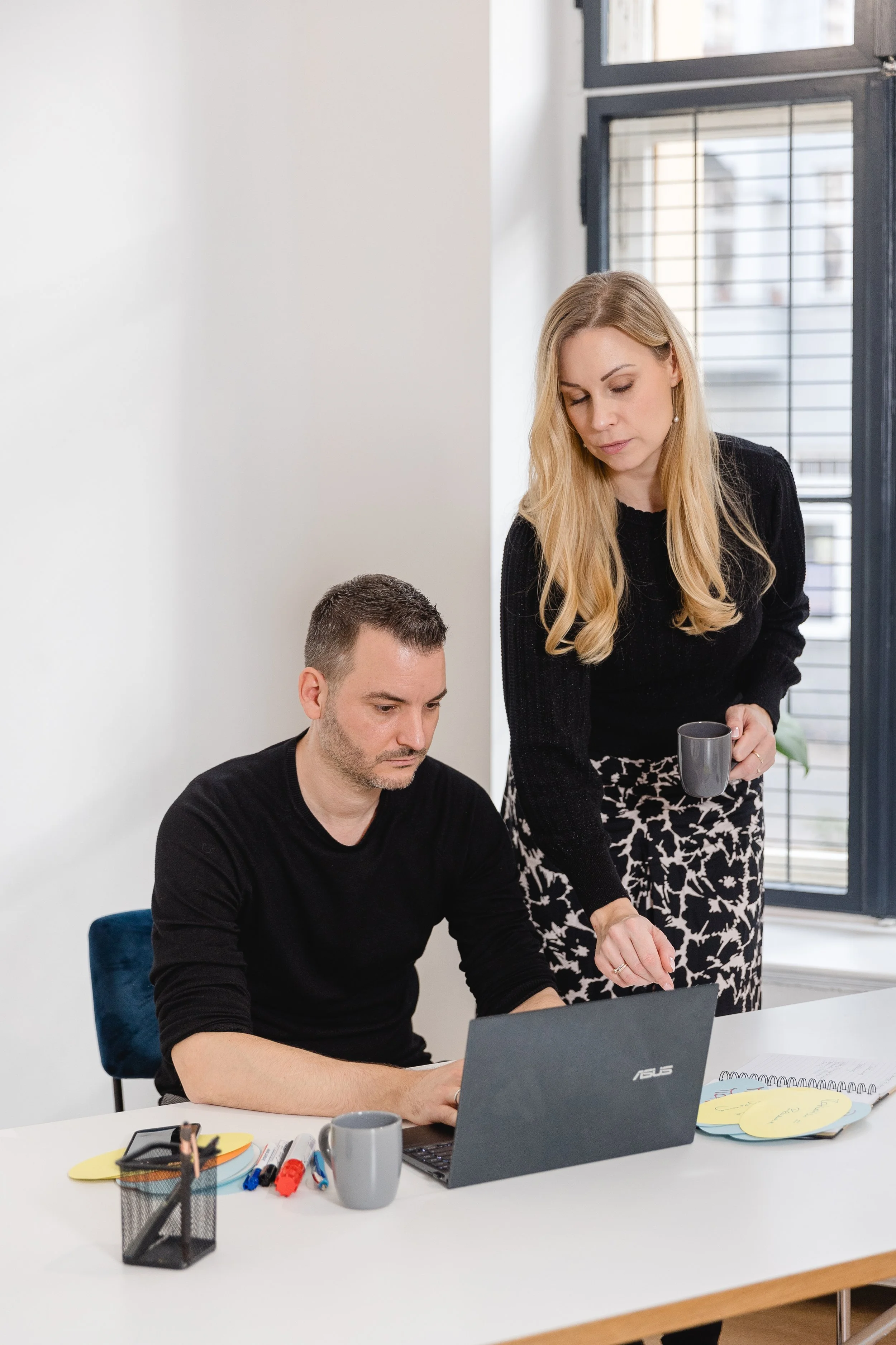Zwei Personen, eine sitzend, und eine stehend, arbeiten zusammen an einem Laptop in einem modernen Büro.