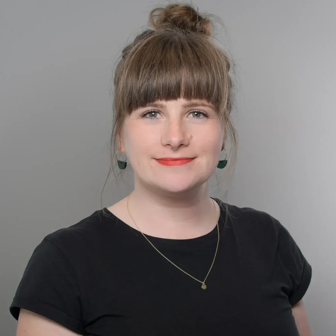 Junge Frau mit braunen Haaren, Dutt, rotem Lippen, in schwarzem T-Shirt, trägt goldene Halskette und grüner Ohrringe, vor grauem Hintergrund.