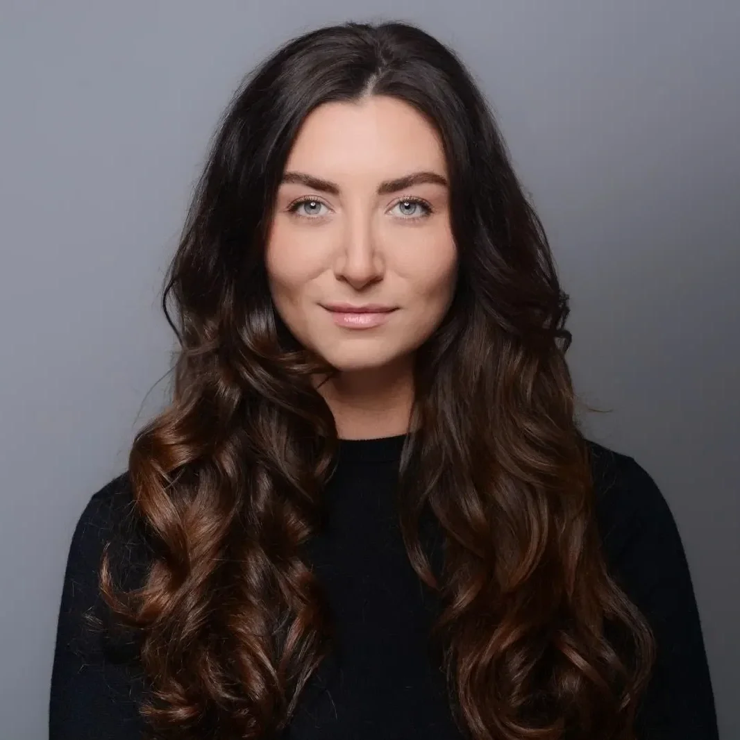 Porträt einer jungen Frau mit langen, braunen, lockigen Haaren, blauen Augen und dezentem Make-up vor grauem Hintergrund.
