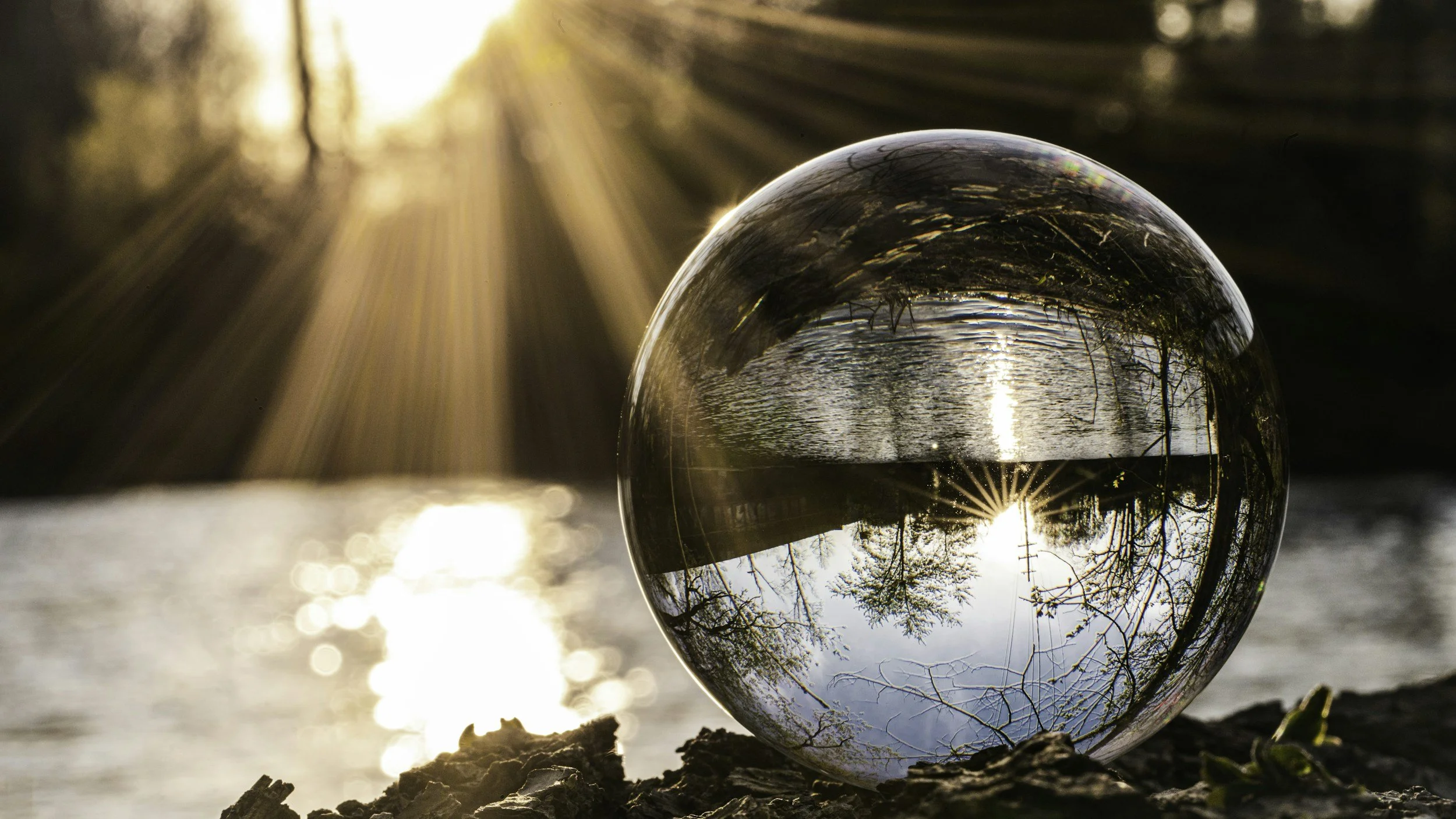 A clear glass sphere reflecting a lakeside scene with trees and a sunset, placed on a rocky surface near water.