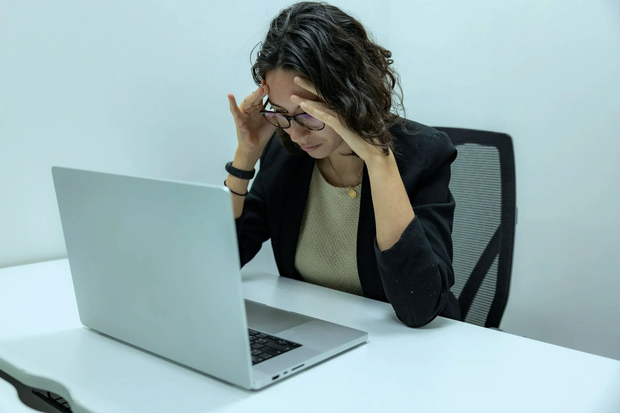 A woman sitting at a white desk, holding her forehead with both hands, looking down at an open silver laptop in front of her, wearing glasses, a black blazer, and a beige top.