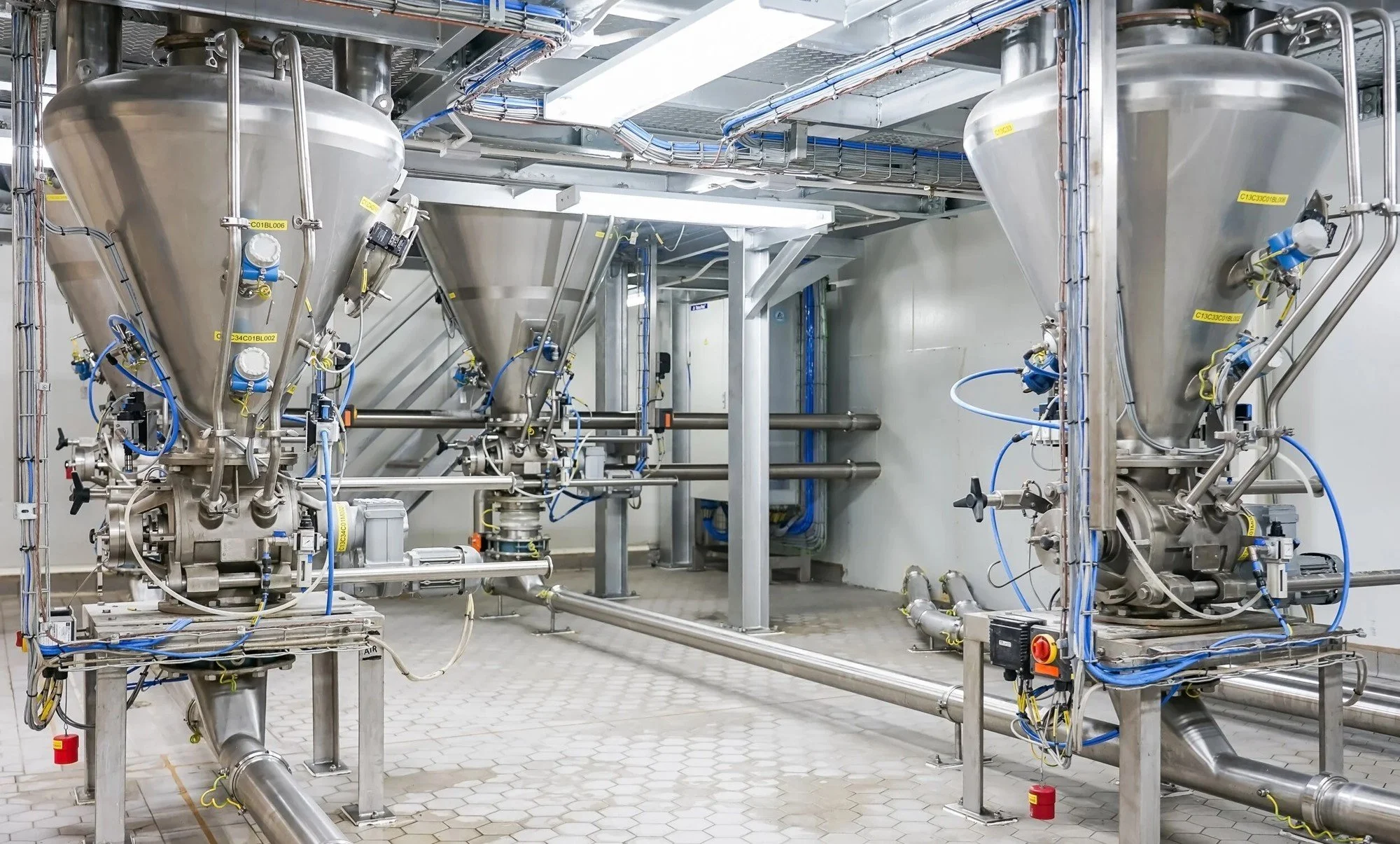 Industrial equipment with large metal funnel-like tanks, pipes, and control valves, set in a clean, well-lit factory or processing plant.