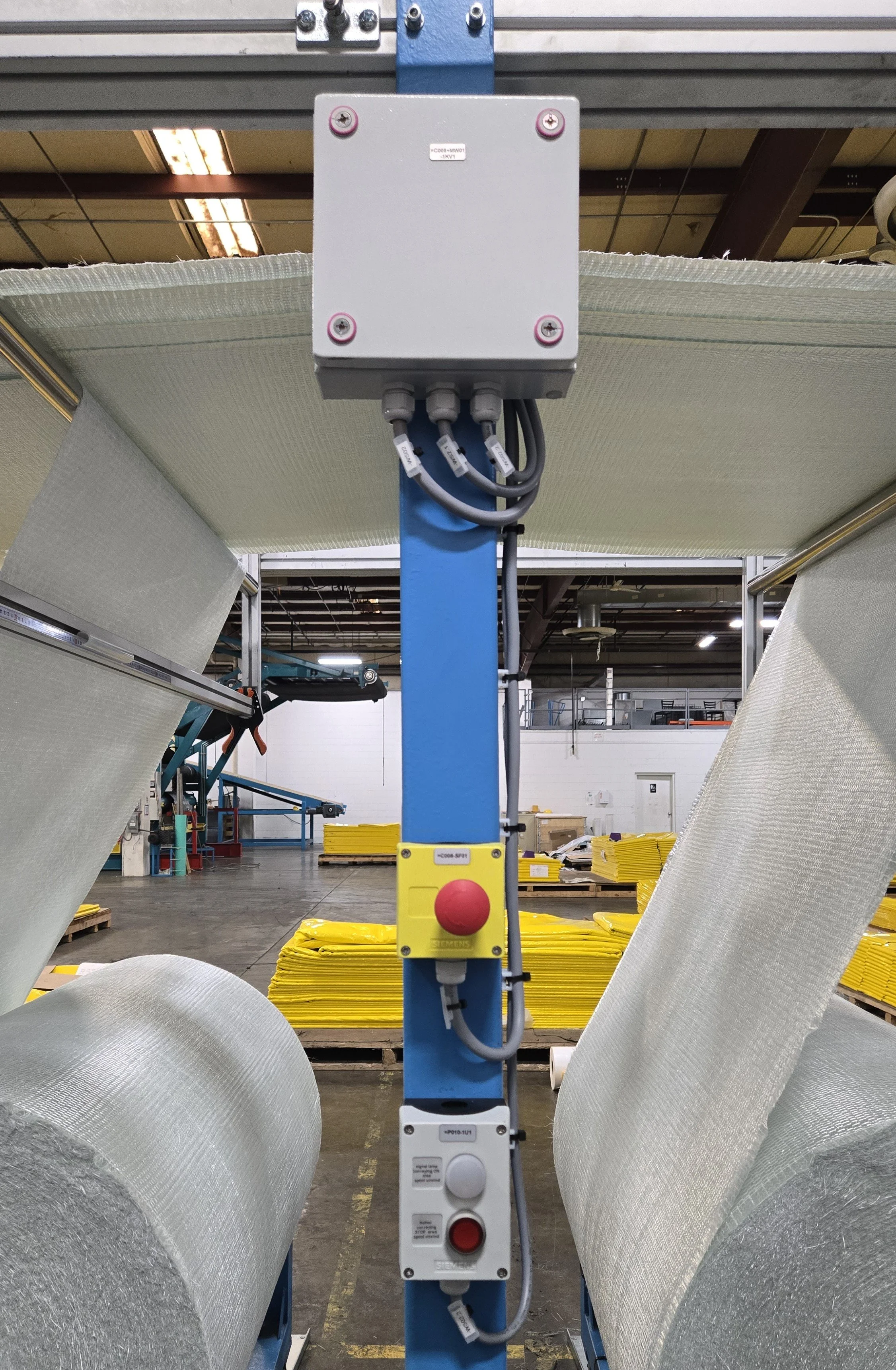 A view of industrial safety control panels mounted on a blue vertical support in a warehouse, with large rolls of reflective insulation material and yellow safety pads in the background.