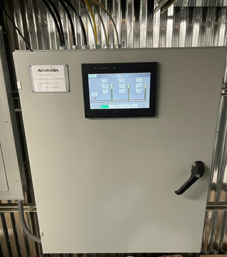Electrical control panel with touchscreen display and a handle, mounted on a metallic wall with exposed wiring above.