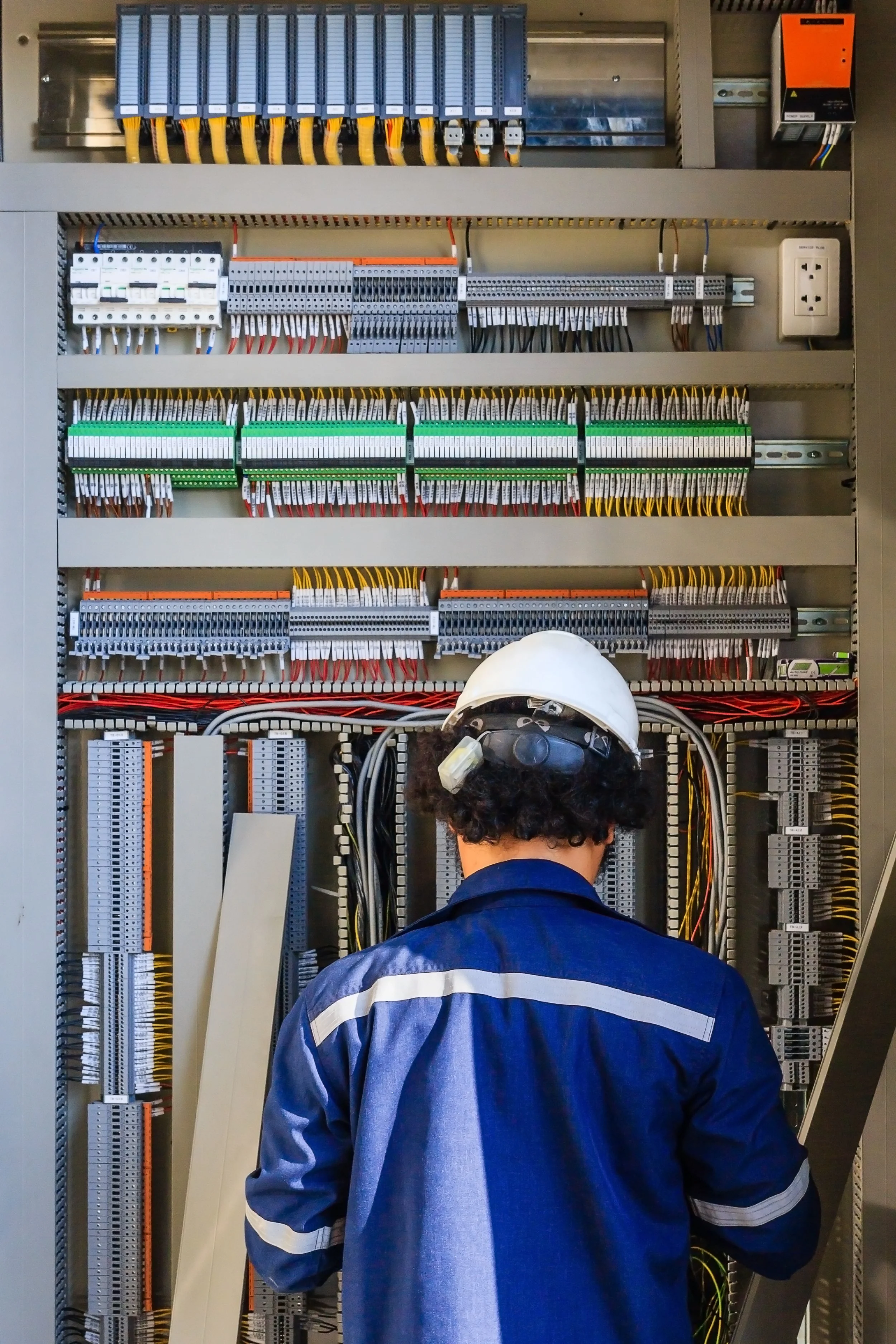 A worker wearing a blue uniform and white safety helmet inspecting electrical control panel with various wiring and circuit components.
