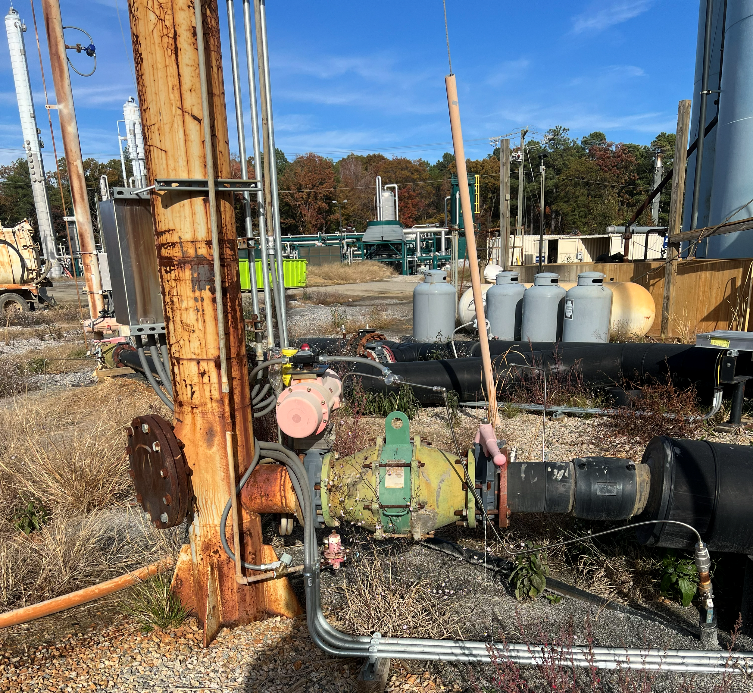 Industrial outdoor scene with rusted metal piping, black insulated pipes, tanks, and electrical equipment on a gravel surface, with trees and blue sky in the background.