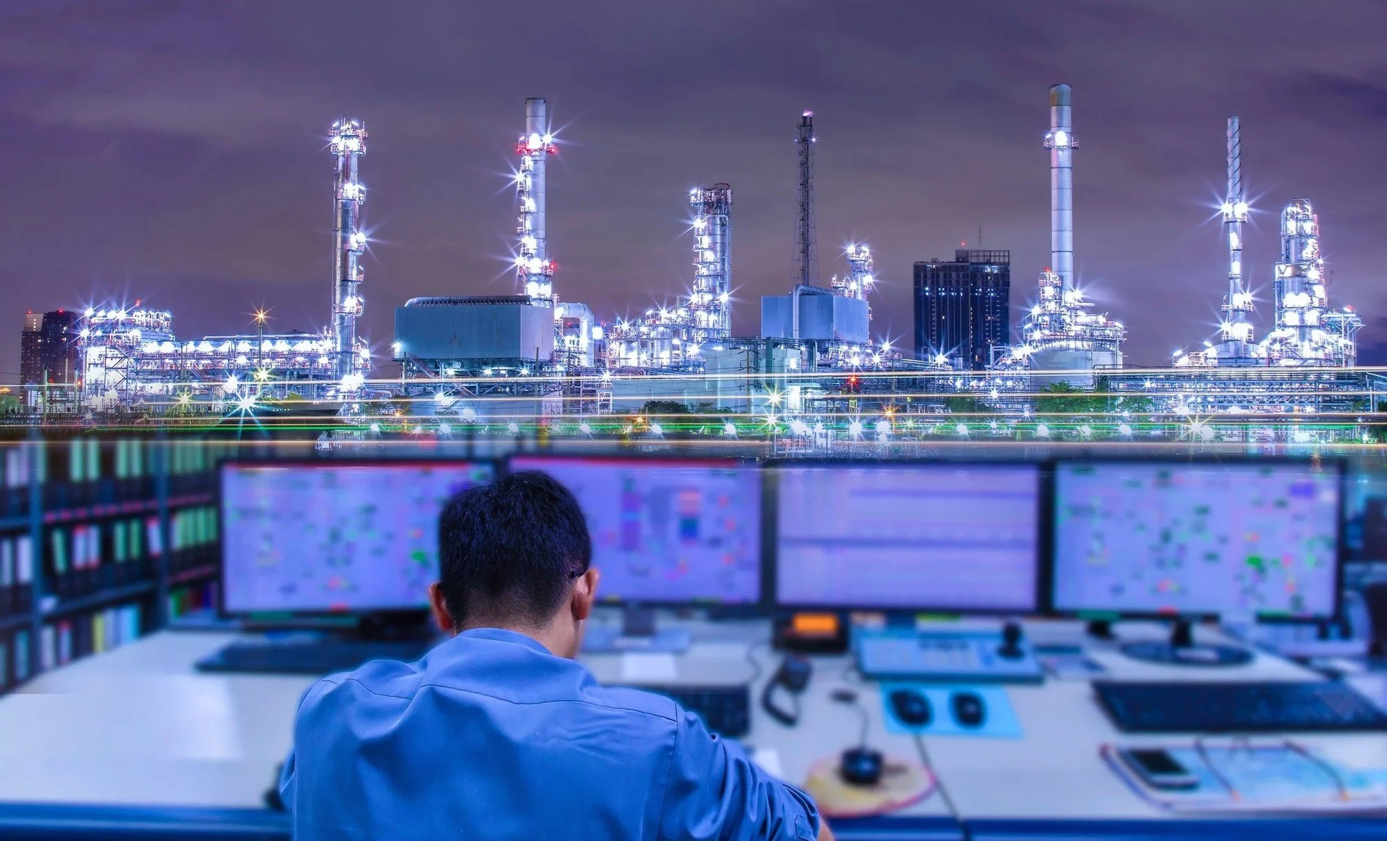 Nighttime scene of a control room with a person monitoring multiple screens, overlooking an illuminated industrial refinery with tall smokestacks.
