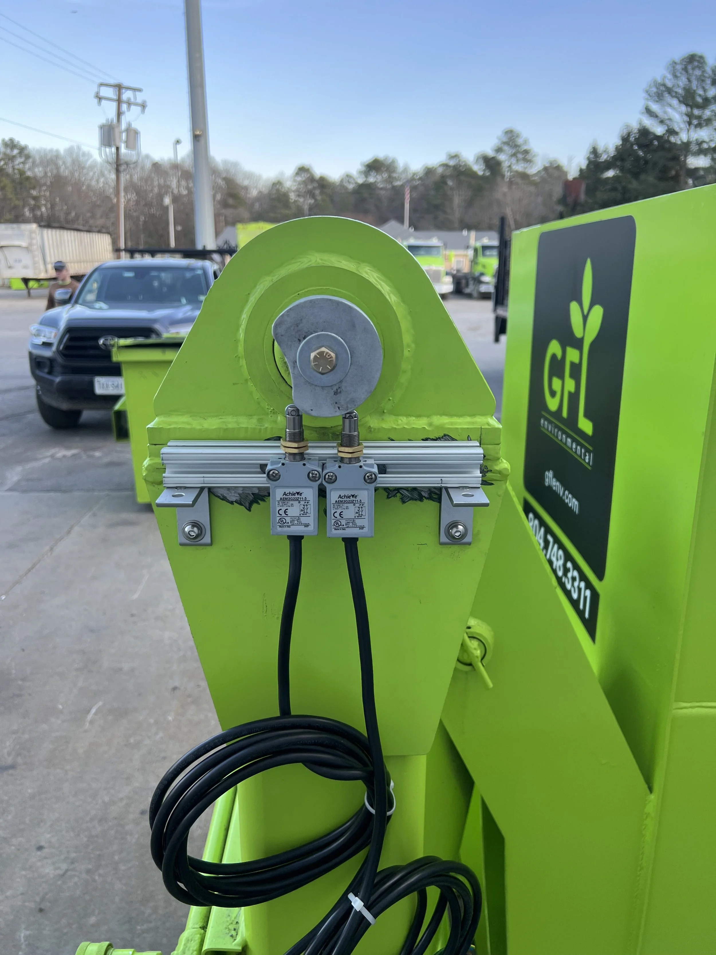 Close-up of a recharging station with electrical cables and components, branded with GFL Environmental, on a parking lot with vehicles in the background.