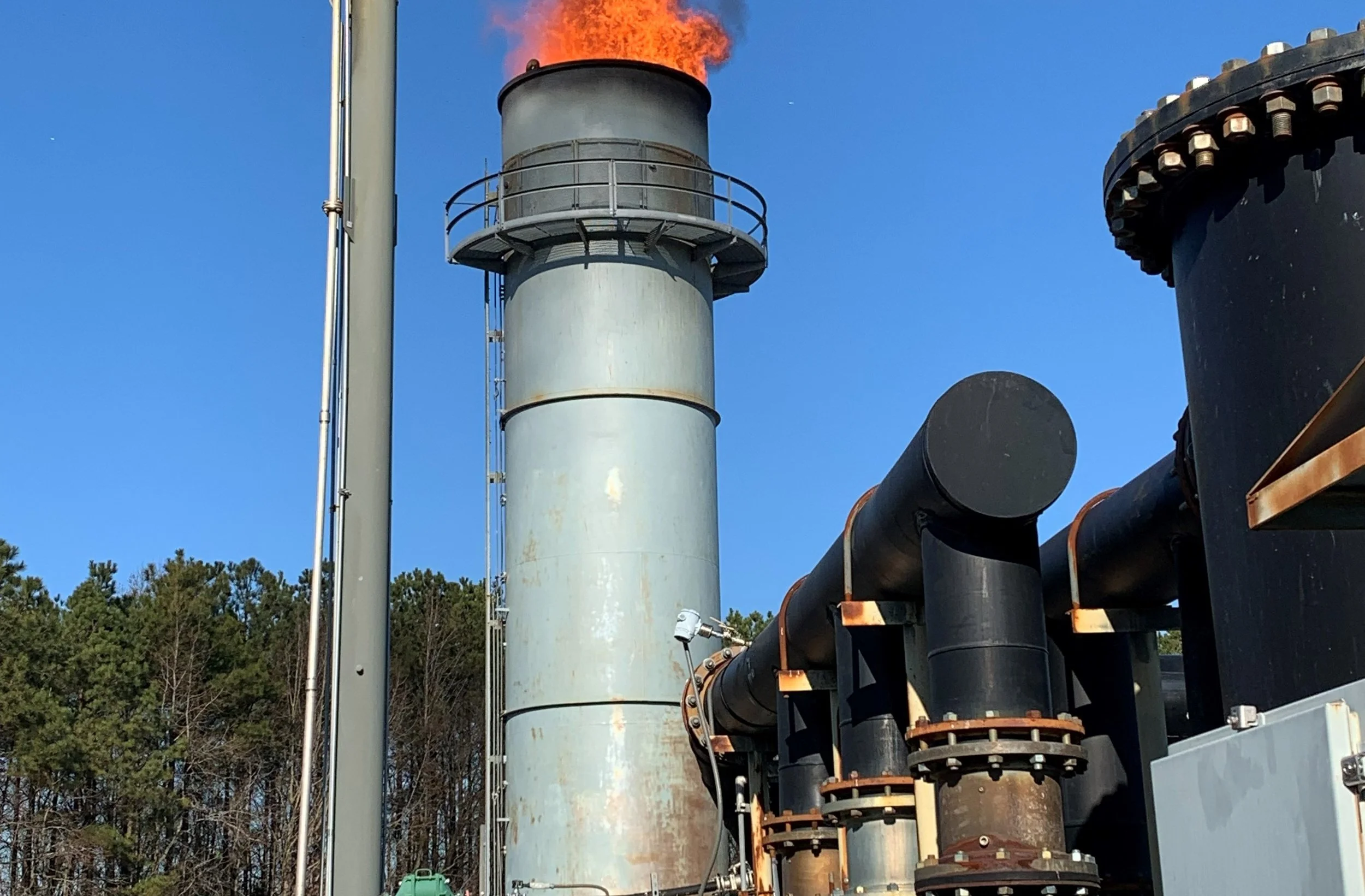 Industrial plant with black pipes, a large gray chimney emitting smoke, and a clear blue sky.