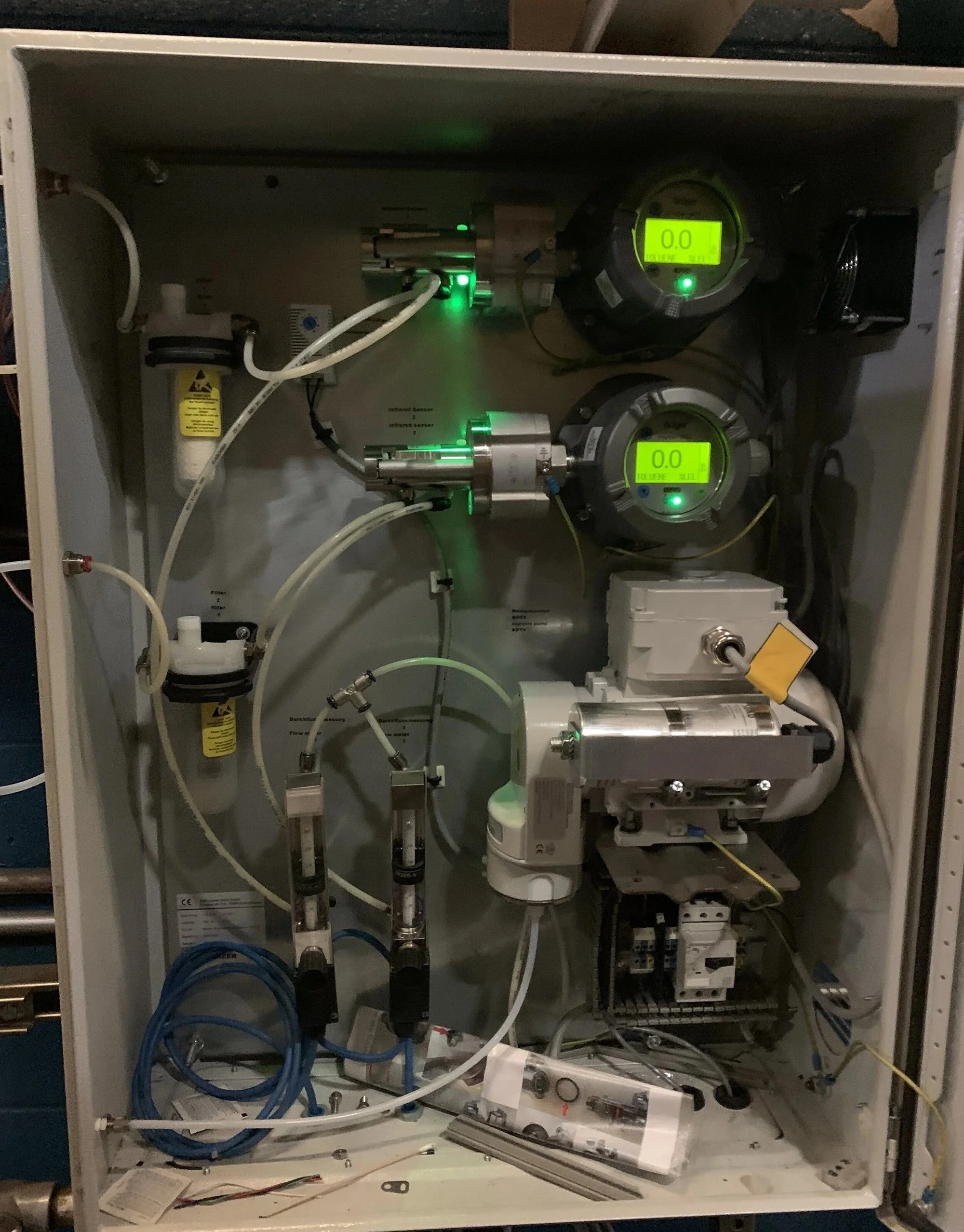 View of an industrial control panel with various gauges, tubes, and electronic components, including glowing green indicators.