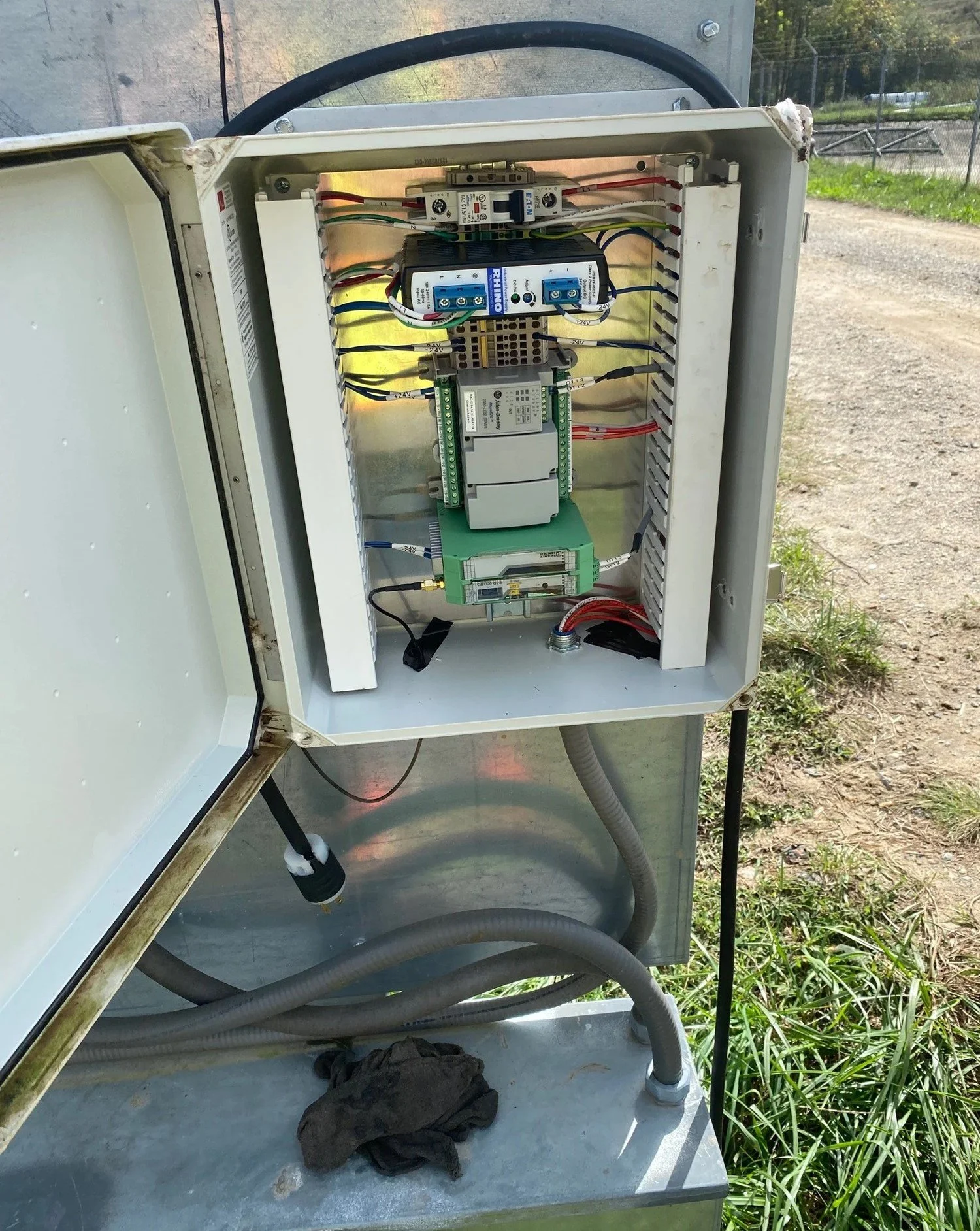 Open electrical control box with various wiring and components inside, mounted outdoors near a grassy area.