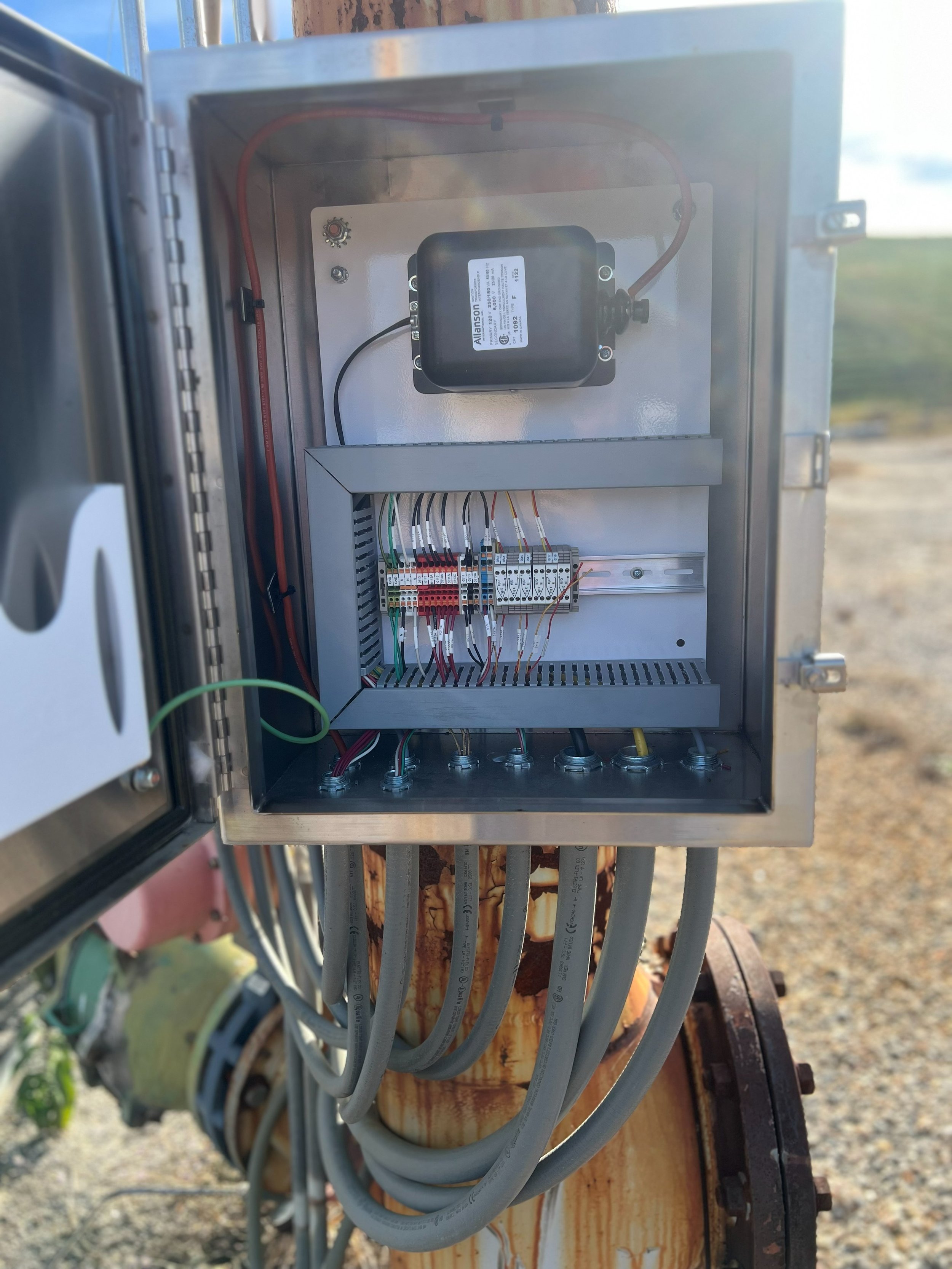 An open electrical control box with wires and circuit components inside, mounted on a rusted metal pole outdoors.
