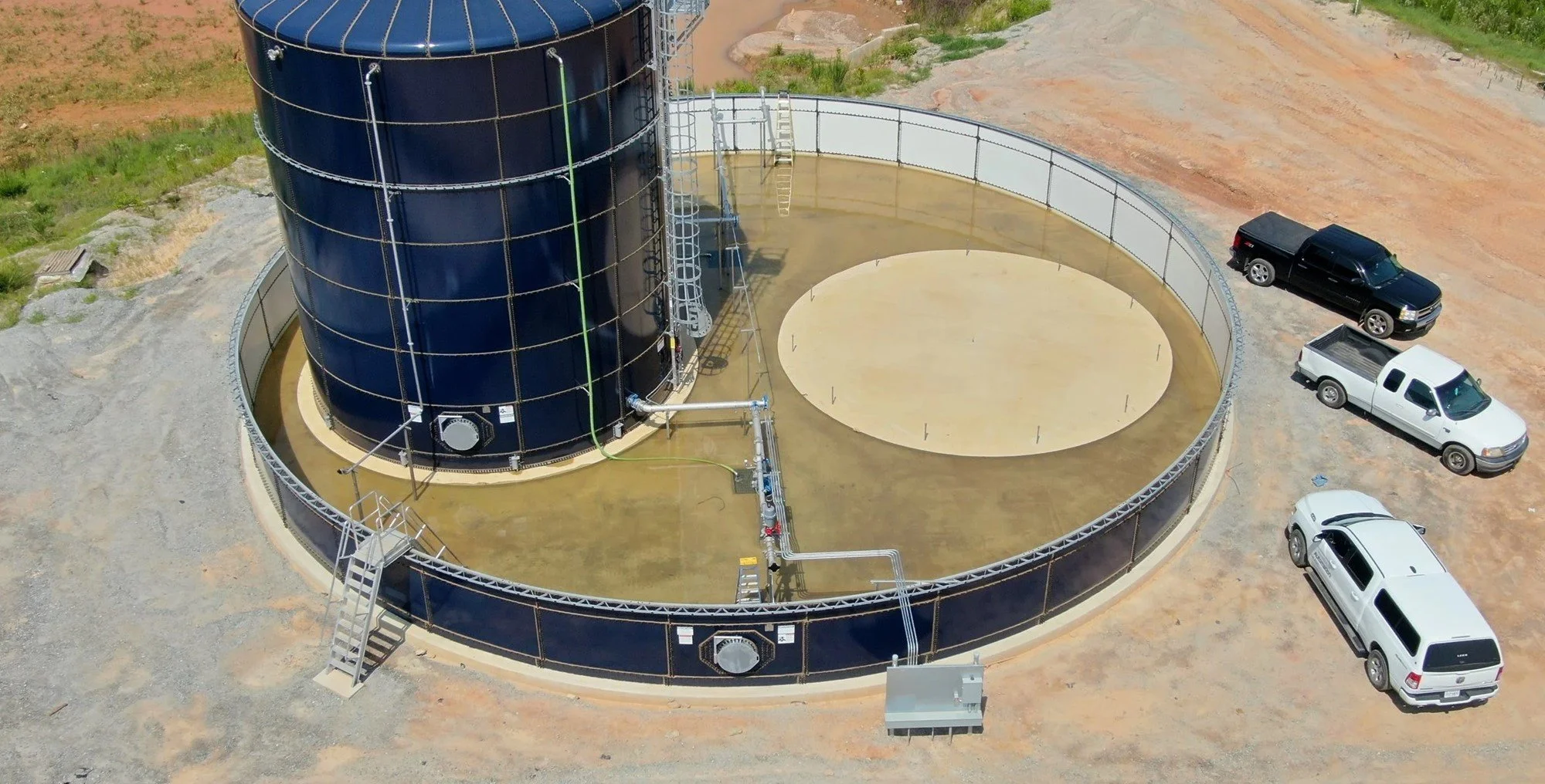 An aerial view of a large circular water tank with a smaller round concrete pad inside the perimeter. Several pipes and ladders are attached to the tank, with three vehicles parked nearby on a dirt area.