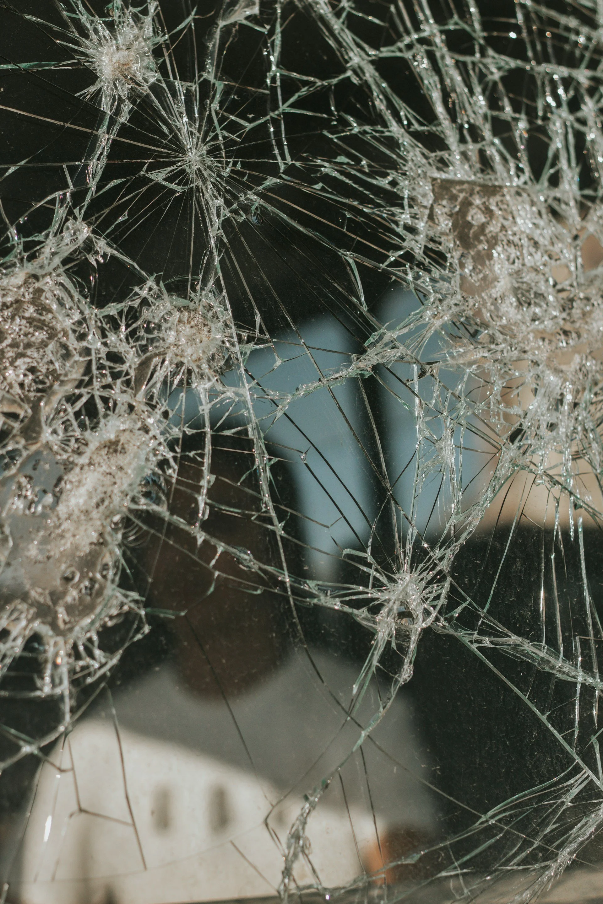 Close-up of shattered glass with intricate cracks spreading outward. A blurred building and sky are visible in the background, conveying chaos.