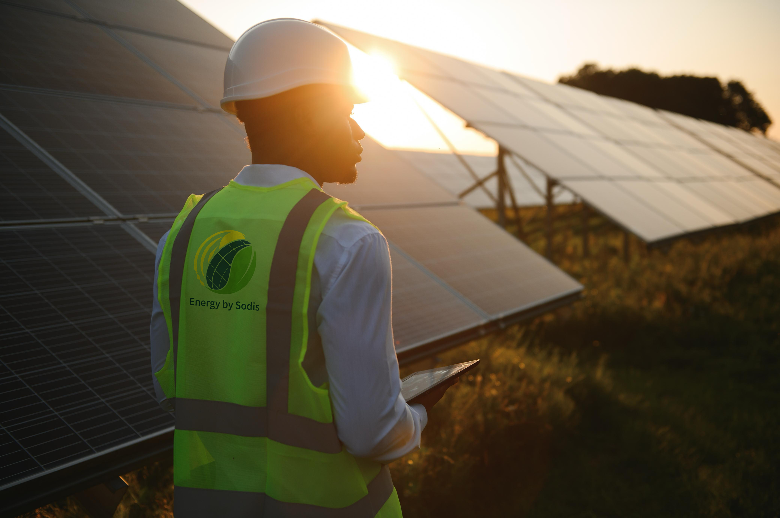 Un homme en gilet de sécurité jaune et casque blanc dans une centrale solaire, utilisant une tablette, avec des panneaux solaires en arrière-plan au coucher du soleil.