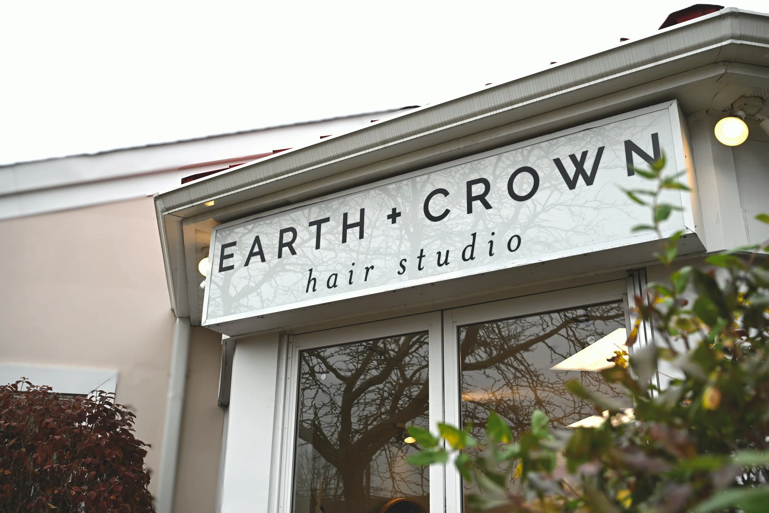 The exterior of a hair studio called 'Earth + Crown.' The sign is white with black text and trees reflecting in the windows below. Welcoming and judgment-free