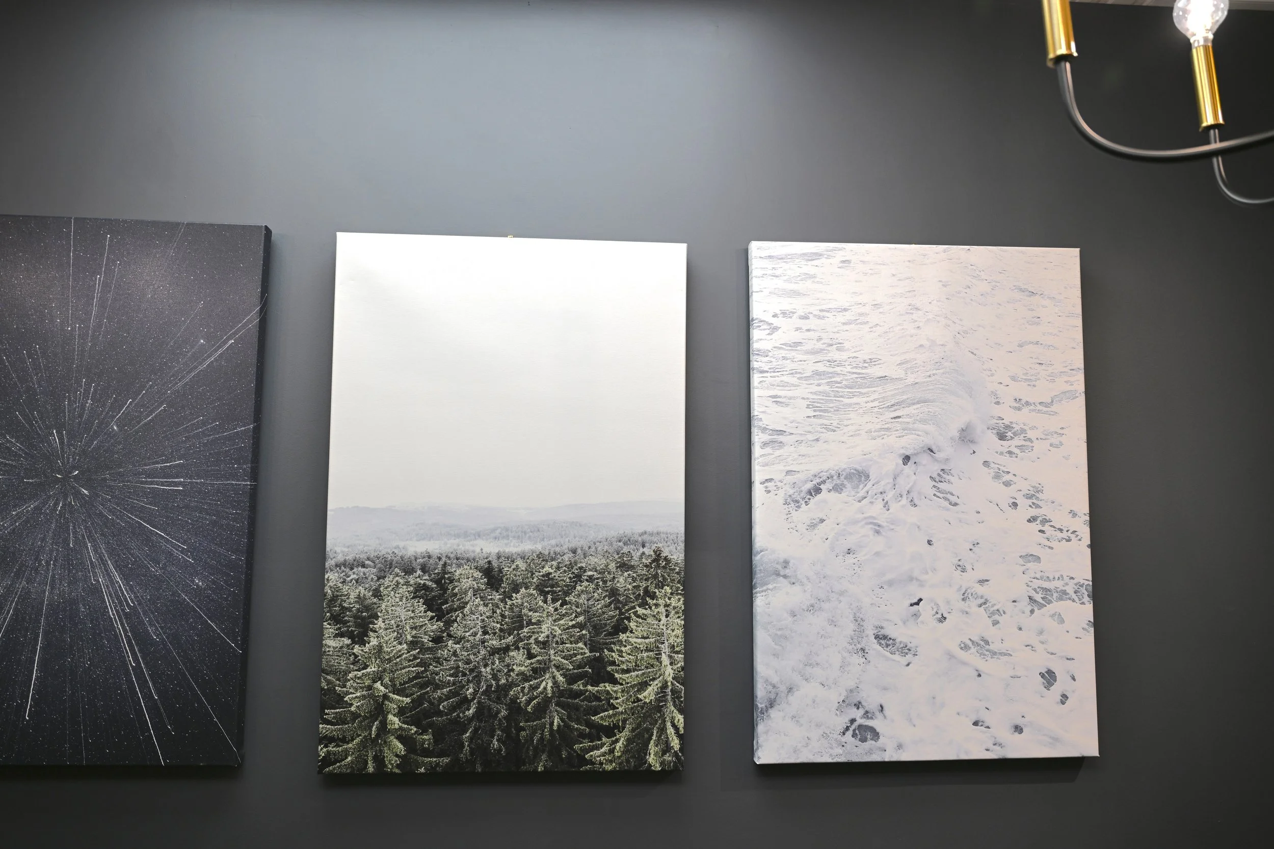 Three landscape photographs on a dark wall. The left photo shows a starry sky with radiating lines. The middle photo depicts a snowy forest with green trees. The right photo captures a white beach scene with foamy waves. Private area and peaceful