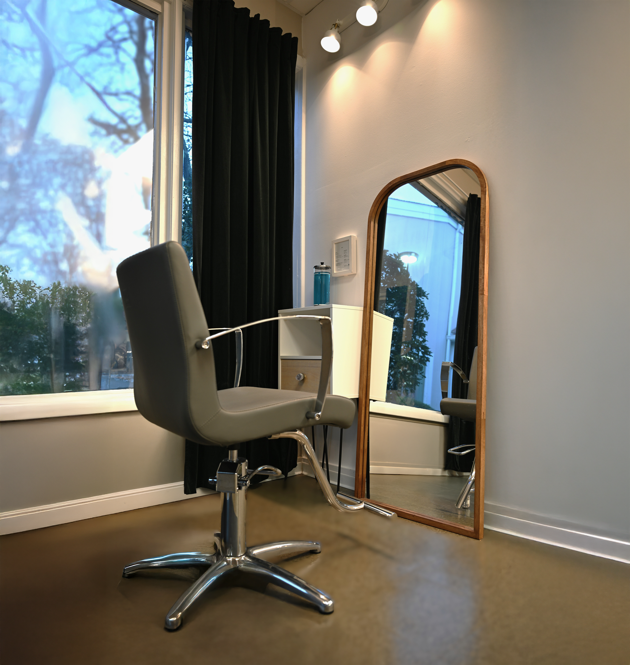 Office space with a gray swivel chair, a large window with blackout curtains, a tall mirror with a wooden frame, and ceiling track lights.