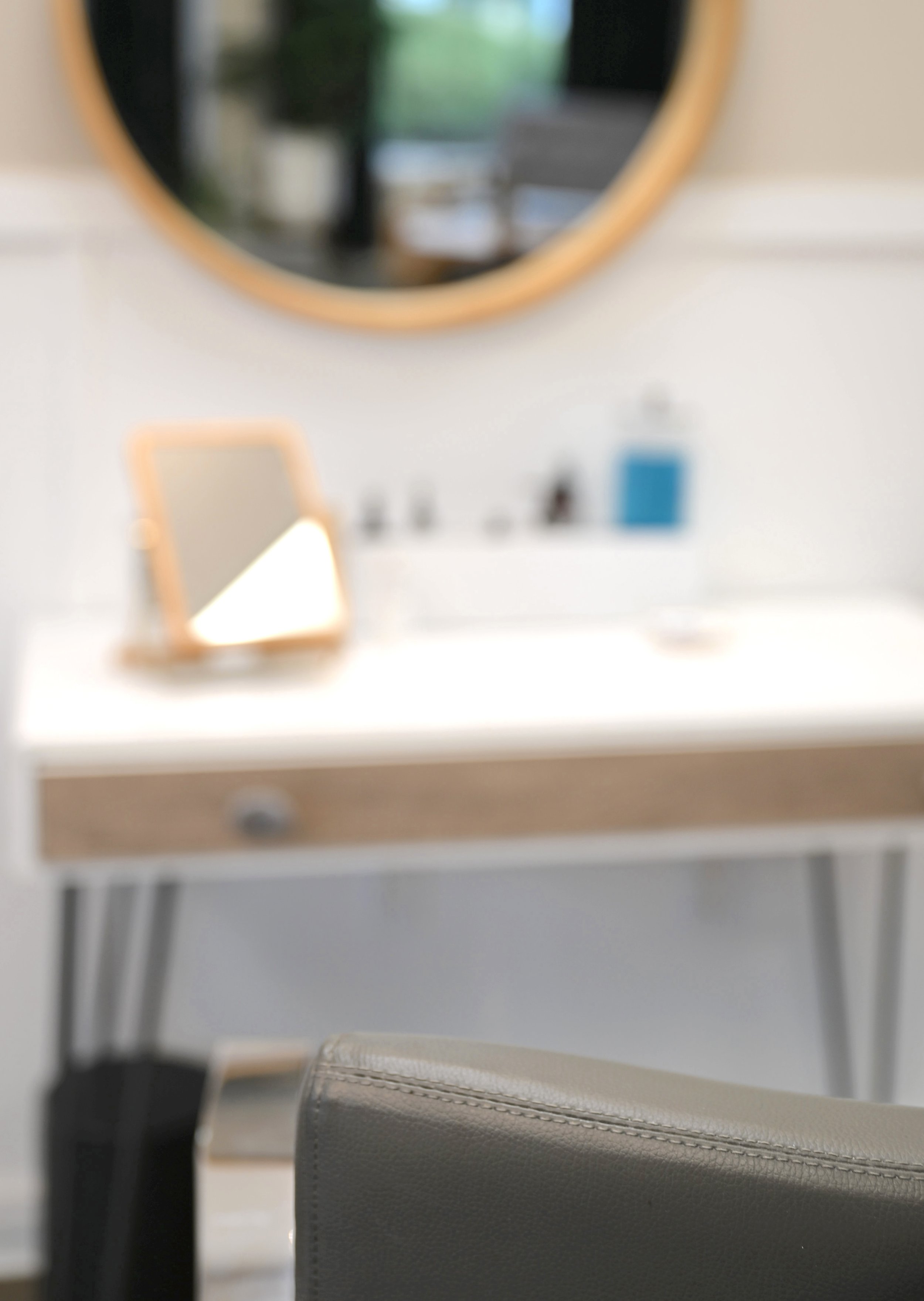 Close-up of a gray chair with black stitching in front of a white table with a small mirror and sanitizer, with a large round mirror on the wall reflecting a blurred outdoor scene. Minimal design for a relaxing hair experience
