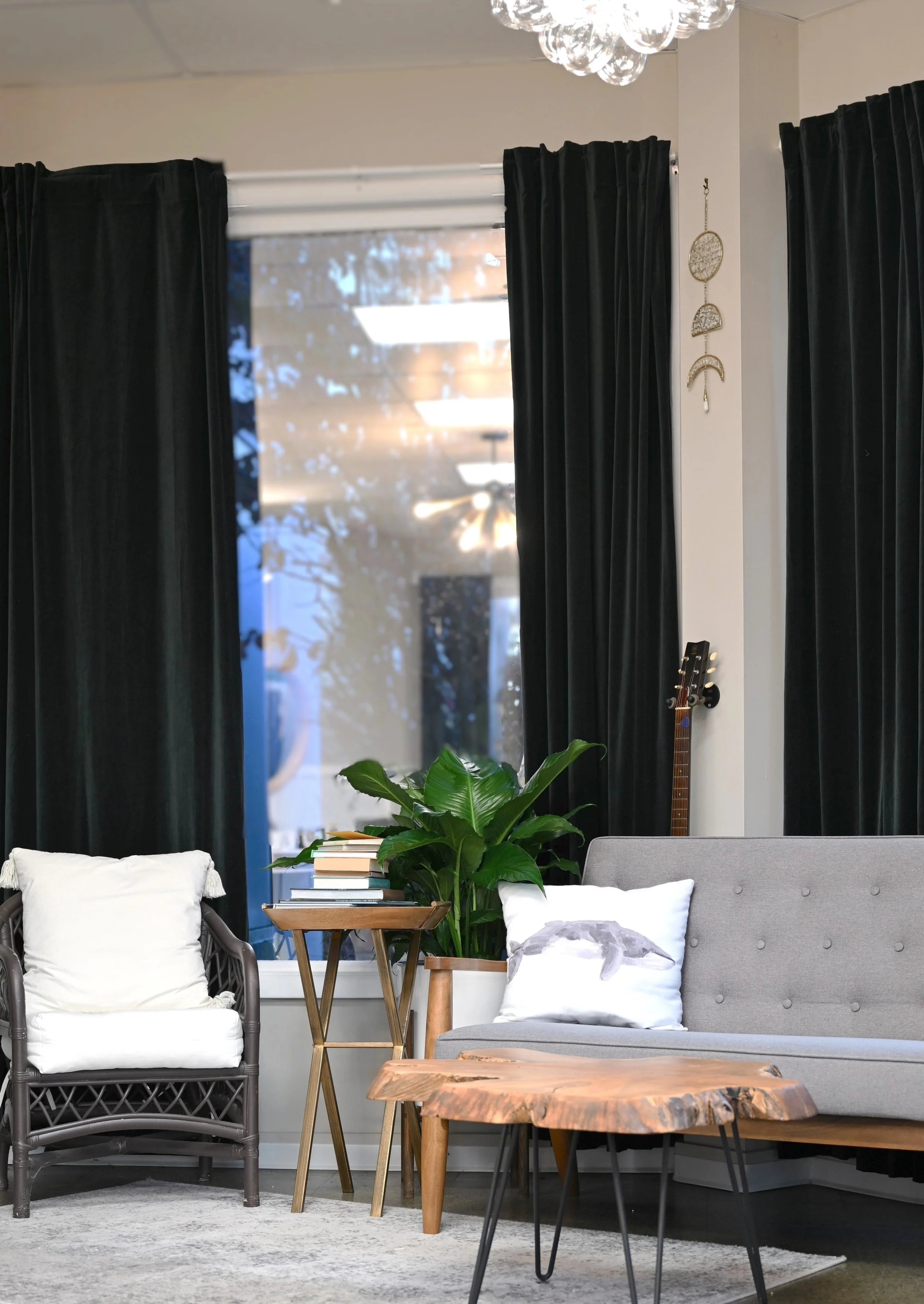 Waiting area black curtains, gray sofa with white pillows, a wicker armchair with white cushion, a wooden coffee table, a small table with stacked books, a large green plant, and a window showing trees outside, with a glass bubble hanging chandelier