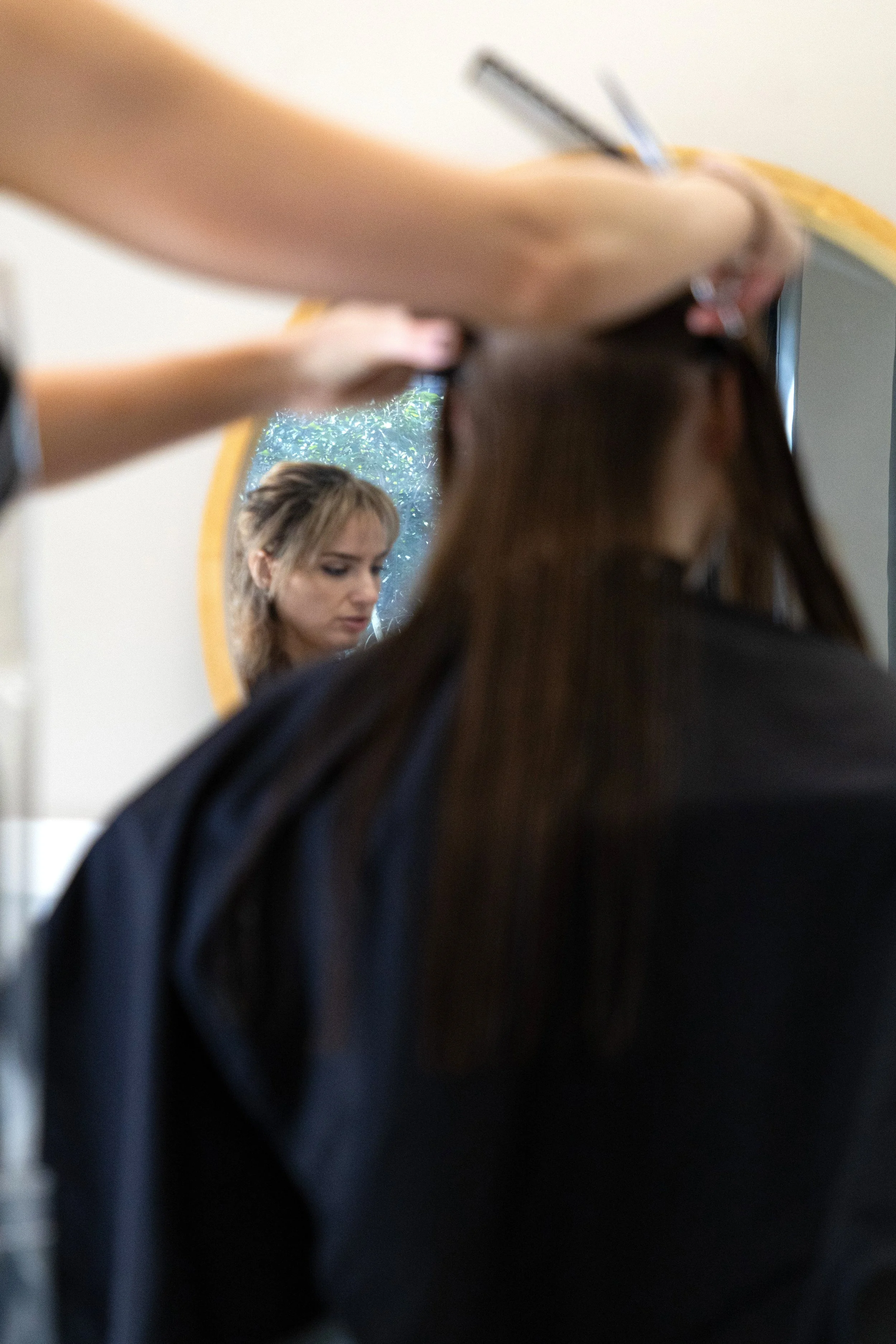A person getting a haircut, with a focus on the mirror showing their reflection and a hairdresser's arm in motion. Professional and quality vegan, low-tox hair services