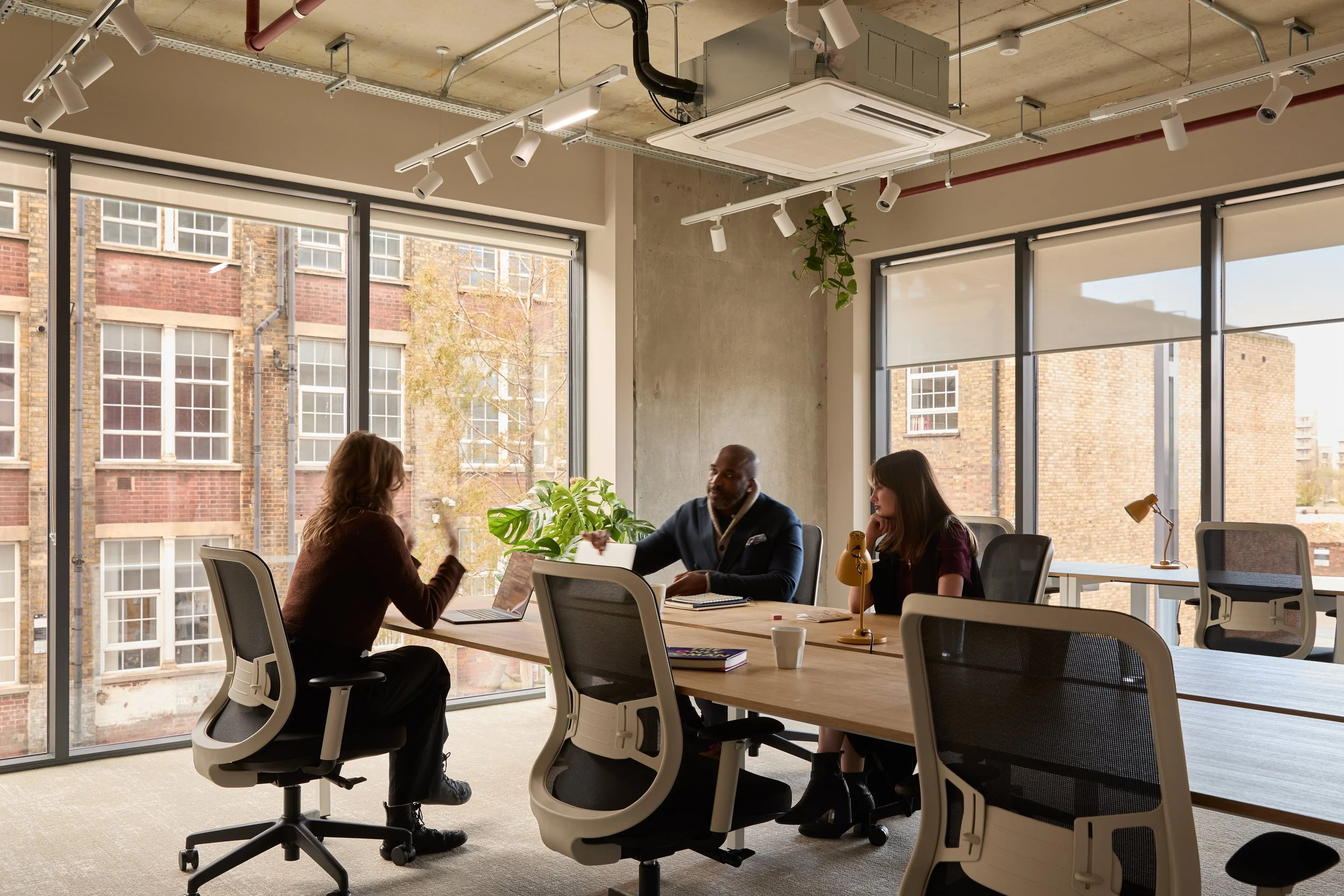 Modern open-plan office with wooden desks, white chairs, computer monitors, hanging pendant lights, pink curtains, and large windows letting in natural light.