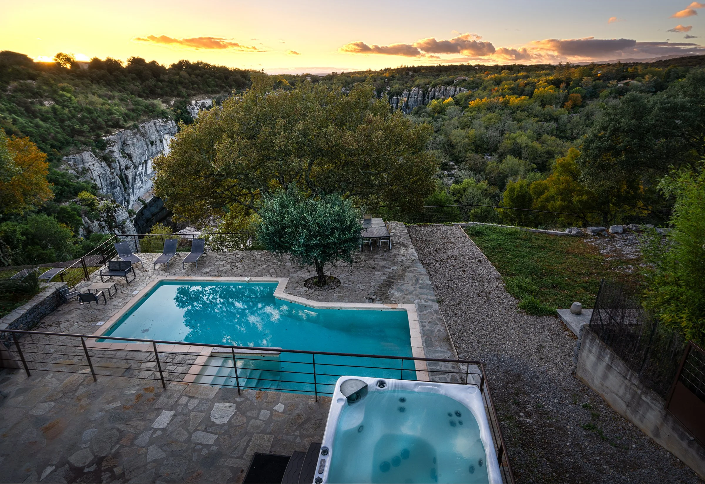 Piscine atypique avec jacuzzi extérieur et vue panoramique sur la nature, falaises et arbres, rivière Beaume coulant au fond, coucher de soleil, ambiance orangée