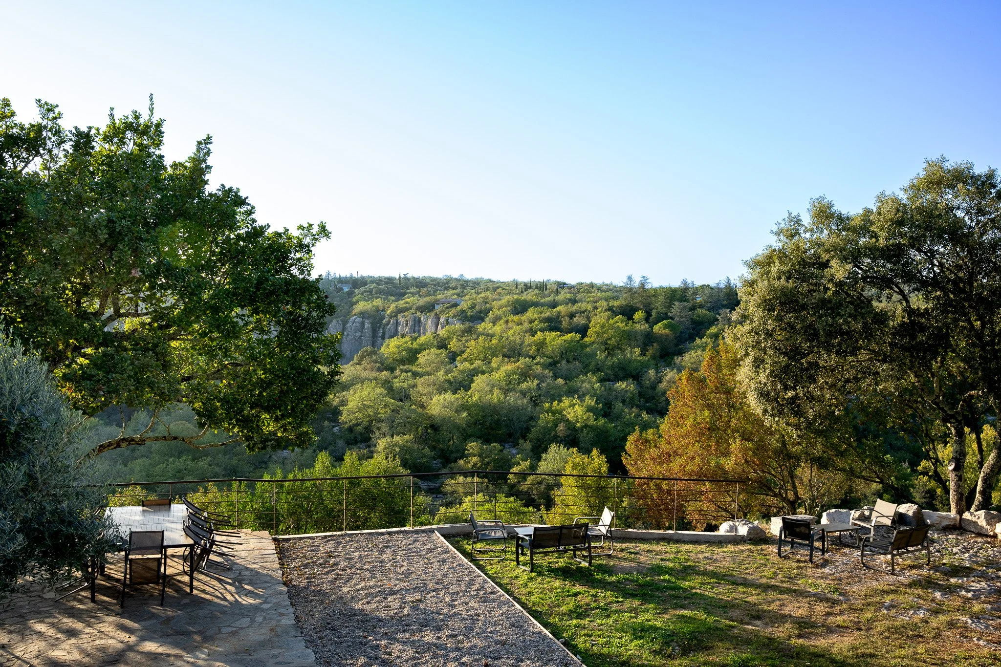 Terrasse avec chaises et fauteuils en métal face à une vallée verdoyante avec des arbres et une falaise en arrière-plan, sous un ciel clair.