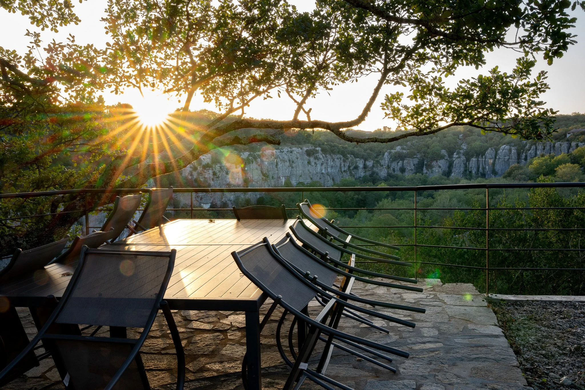 Une terrasse avec une table et des chaises en métal, accueil grande capacité, surplombant une vallée avec des falaises rocheuses, sous un soleil couchant ou levant à travers les branches d'un chêne centenaire.