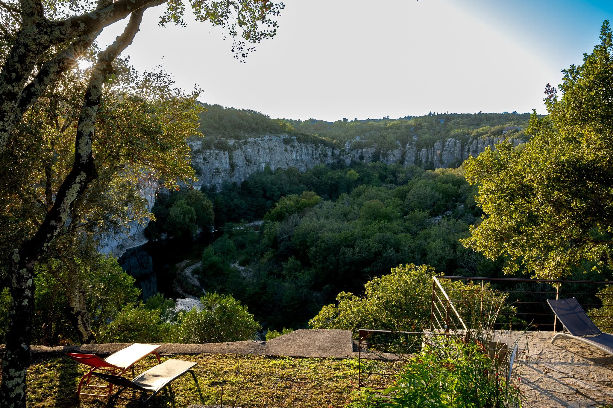 Une vue d'une vallée verdoyante avec des falaises blanches, des arbres luxuriants, des chaises longues sur une terrasse ensoleillée.