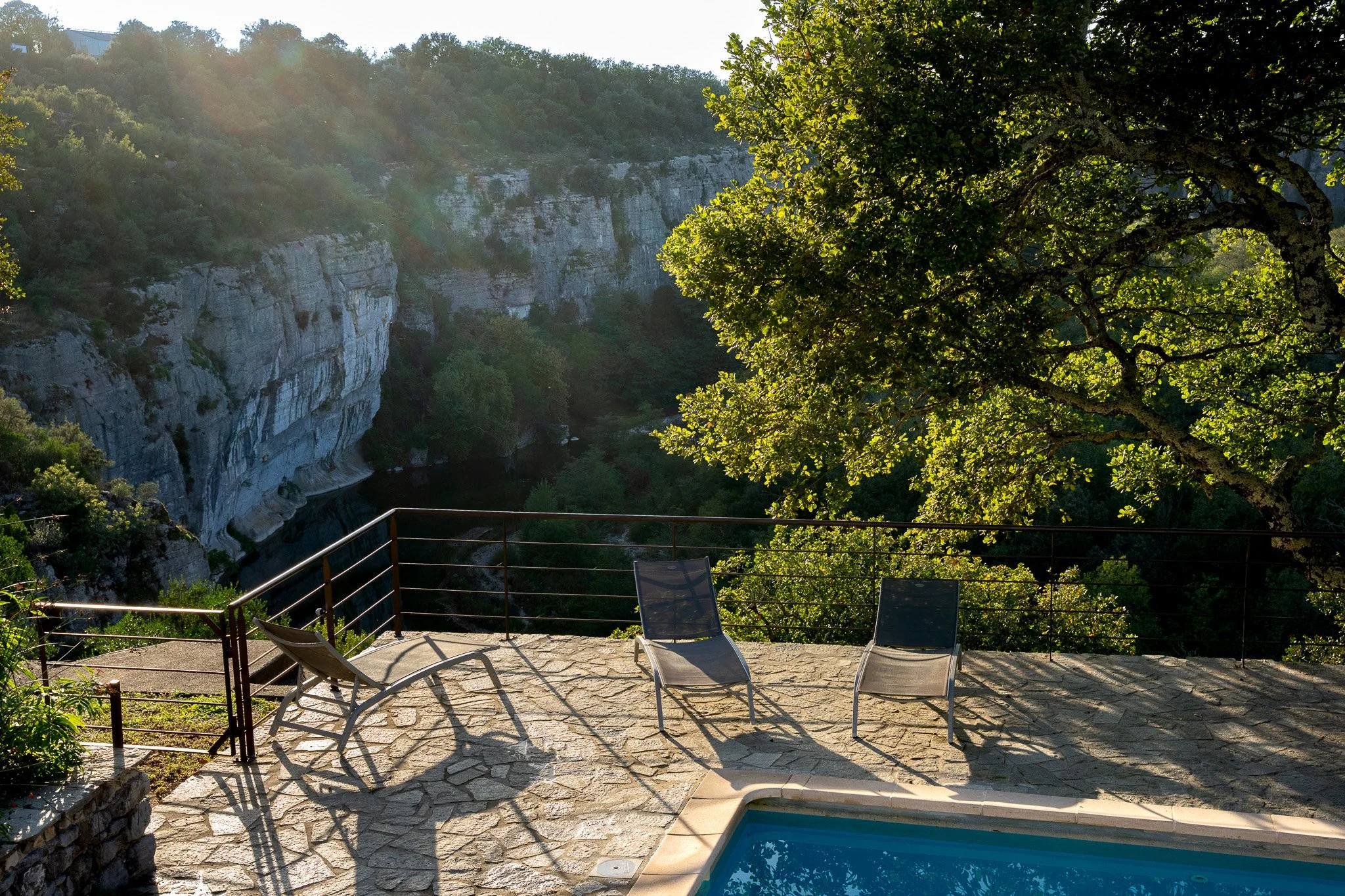 Une terrasse avec un bassin de piscine, deux chaises longues et un fauteuil, donnant sur un canyon avec des falaises rocheuses, entouré d'arbres verts et sous un ciel ensoleillé; barrières de sécurité autour de la piscine. 