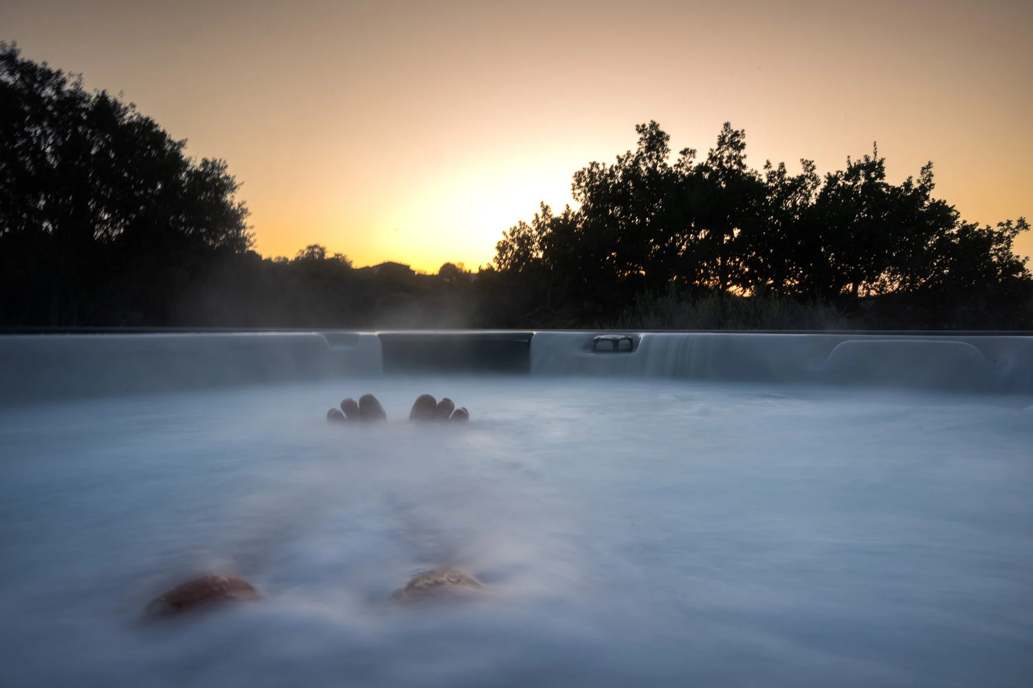 Personnes se relaxant dans un jacuzzi extérieur au coucher du soleil, des arbres en silhouette en arrière-plan.