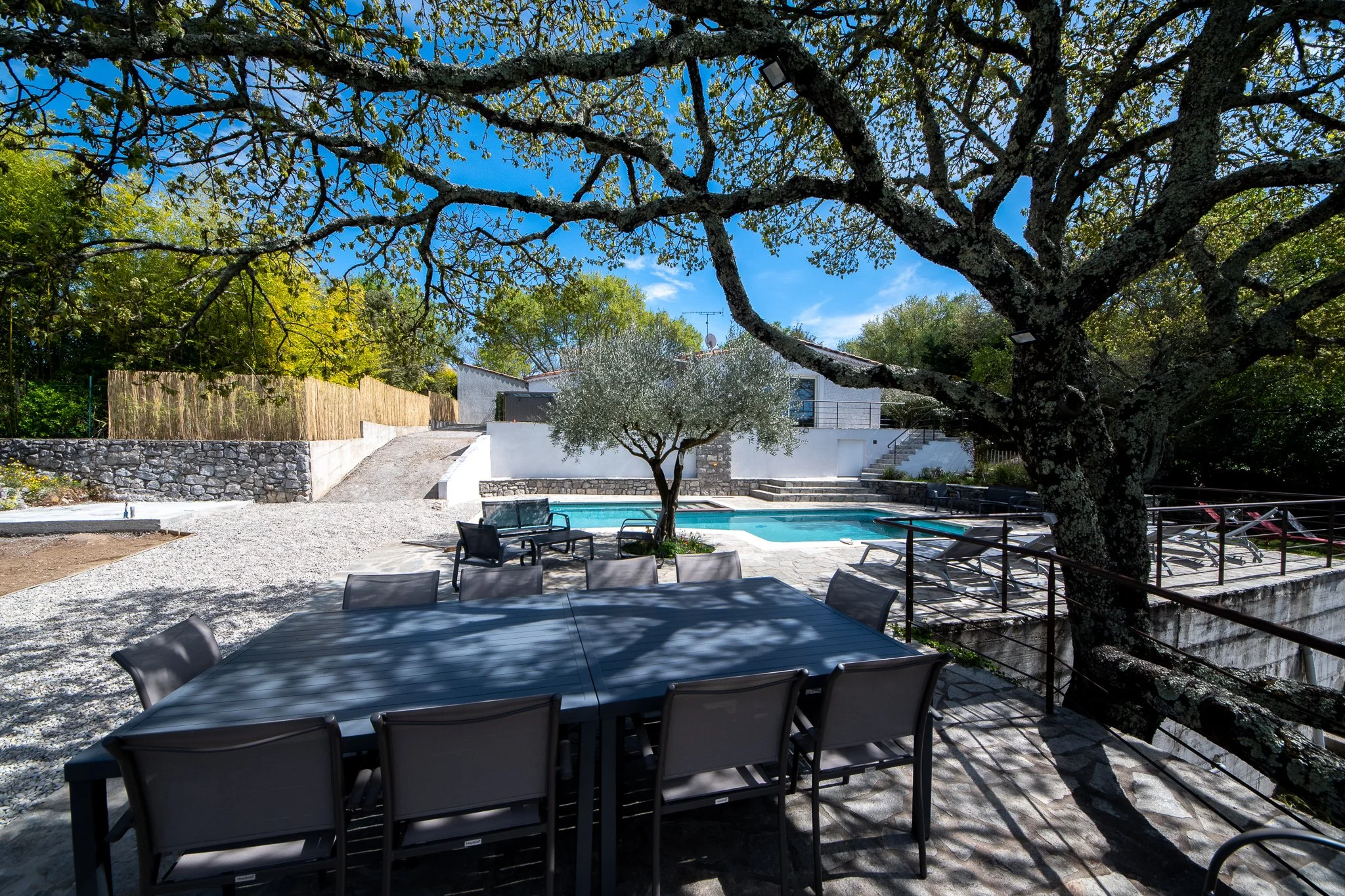 Une terrasse avec une grande table entourée de chaises, ombragée par un grand arbre. En arrière-plan, une piscine et un olivier, et le gîte, avec un patio en pierre et une clôture en bois et en pierre. Le ciel est bleu avec quelques nuages.