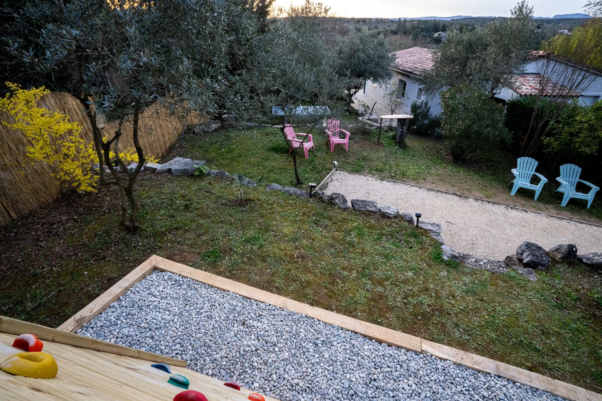 Vue du jardin avec plusieurs chaises de couleur (rose, bleu) disposées autour d'une petite zone de gravier, arbres, un espace vert avec de la végétation et une barrière en bambou