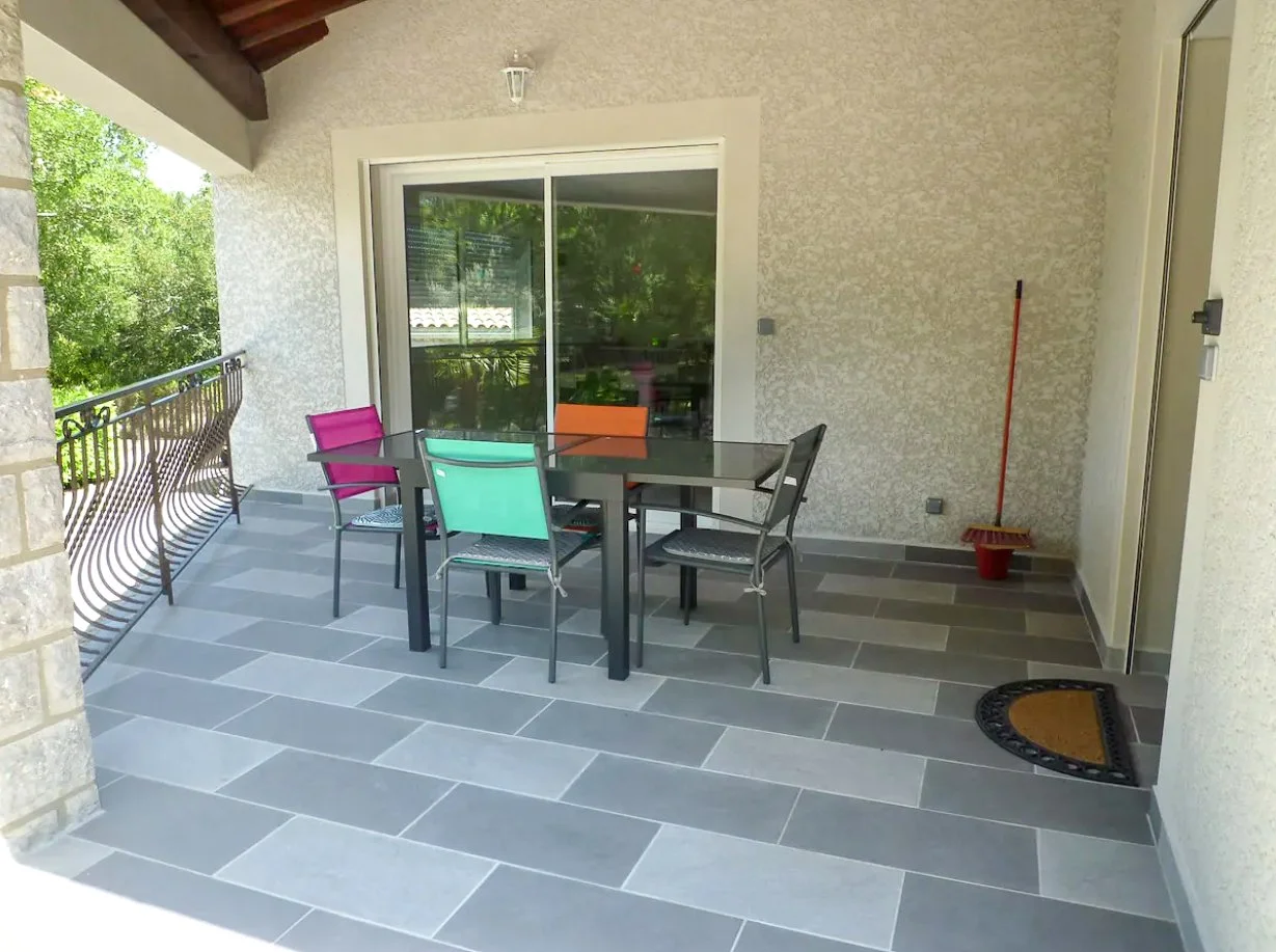 Balcon avec table et chaises colorées, balustrade en métal, porte-fenêtre, balconnet entouré de murs en pierre et mur généralement beige avec un balai et un paillasson au fond.