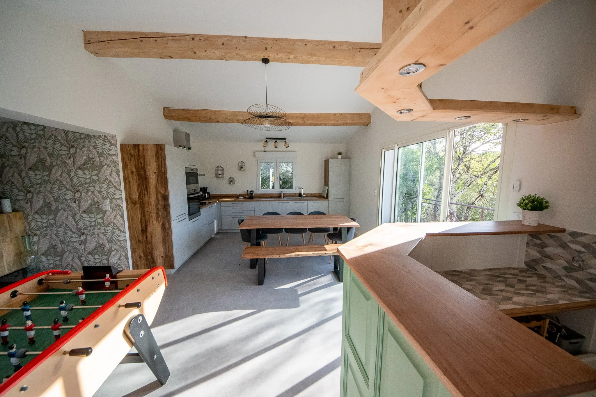 Cuisine lumineuse avec plafond en bois, grande fenêtre, table en bois avec 4 chaises, mur d'accent avec papier peint à motif de feuilles, baby-foot, bar, porte vitrée ouvrant sur la terrasse, la piscine et la vue panoramique.