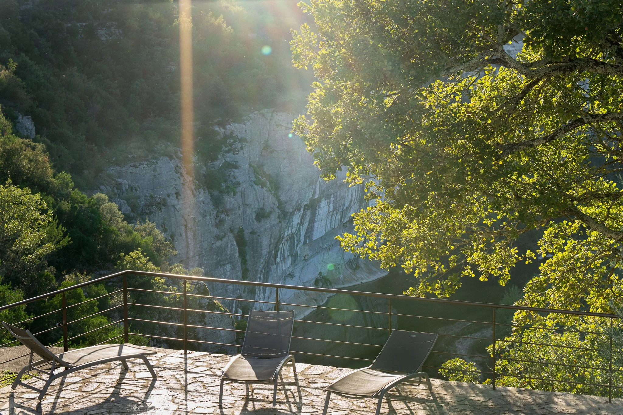 Chaises longues sur une terrasse en pierre avec vue sur une rivière et des falaises, ensoleillée avec des rayons de soleil filtrant à travers les arbres.