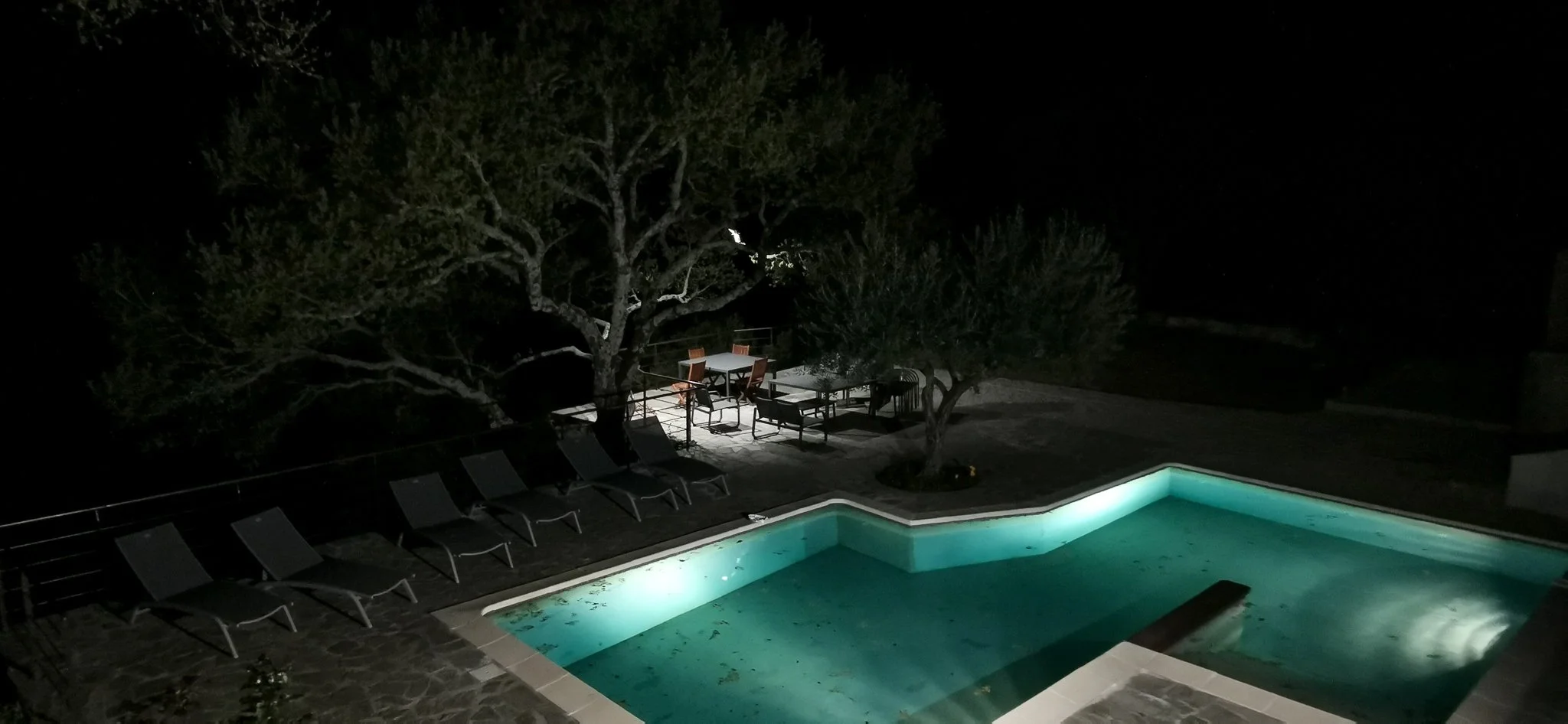 Piscine éclairée la nuit avec des chaises longues autour, arbres et mobilier de jardin en arrière-plan