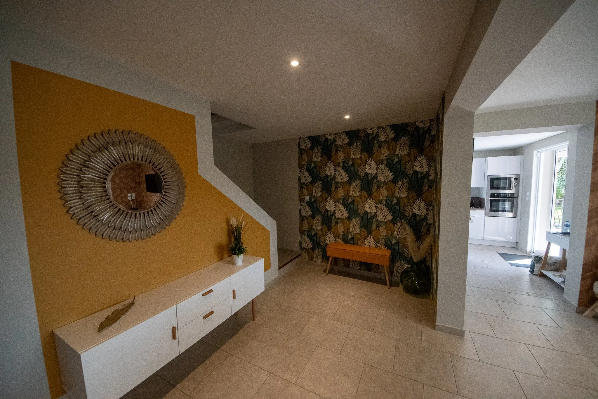 Intérieur d'une maison avec un mur jaune et un miroir décoratif, un meuble blanc avec une plante, et un mur avec un papier peint floral. En arrière-plan, une cuisine visible avec des appareils en acier inoxydable et une porte donnant sur l'extérieur.