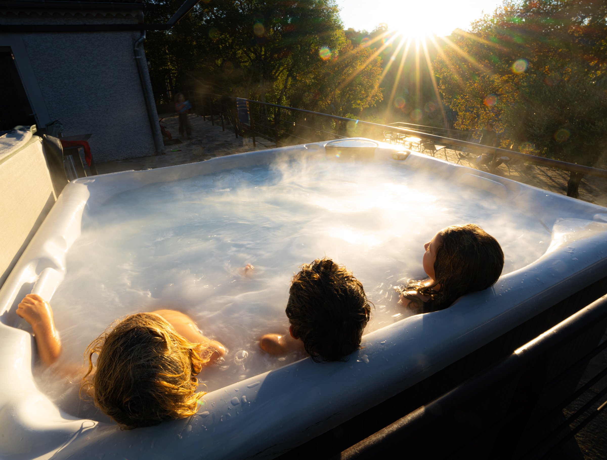 Trois enfants dans un jacuzzi extérieur au coucher du soleil, entourés d'arbres aux feuilles d'automne.