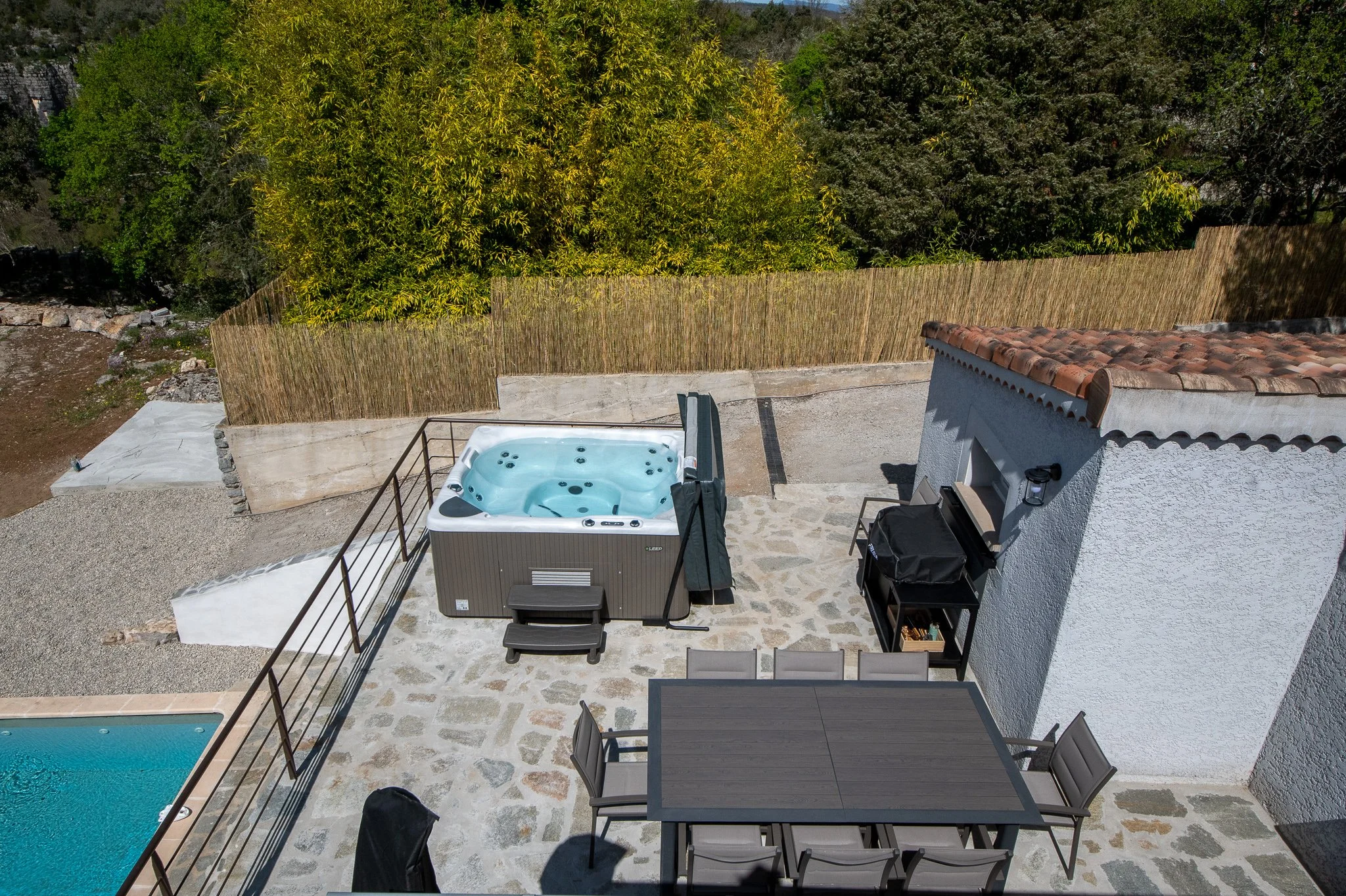 Vue aérienne d'une terrasse avec jacuzzi, table avec six chaises, barbecue, et une piscine à côté. Bordure en pierre, clôture en bambou et arbres en arrière-plan.