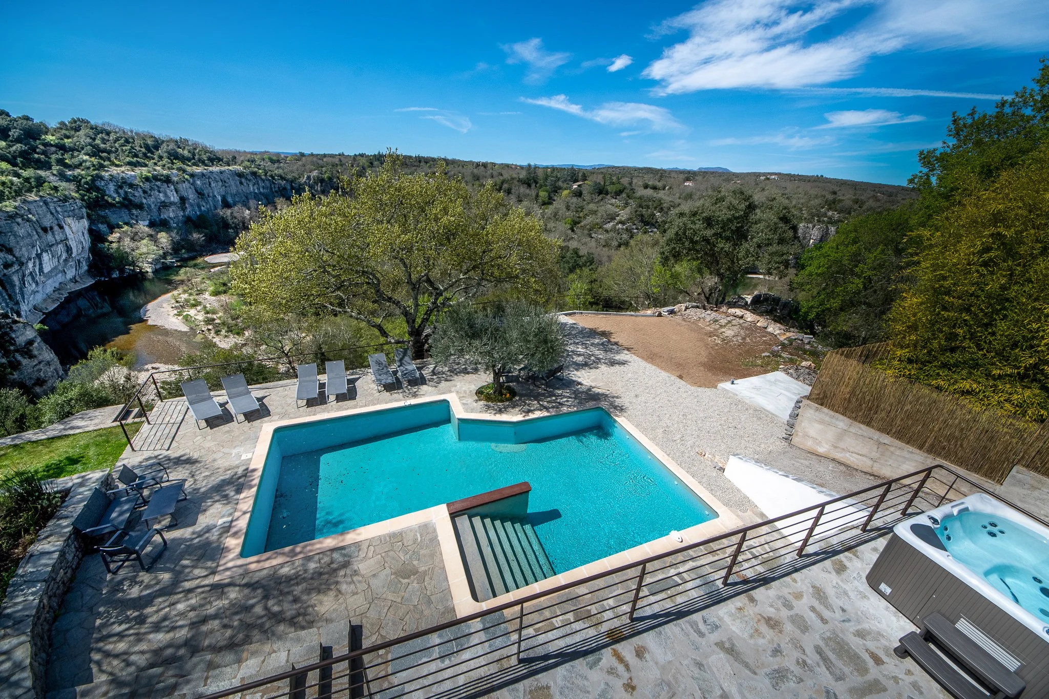 Piscine avec vue panoramique sur la rivière Beaume, les falaises et les arbres, chaises longues, arbre, jacuzzi en extérieur, ciel bleu et paysage naturel.