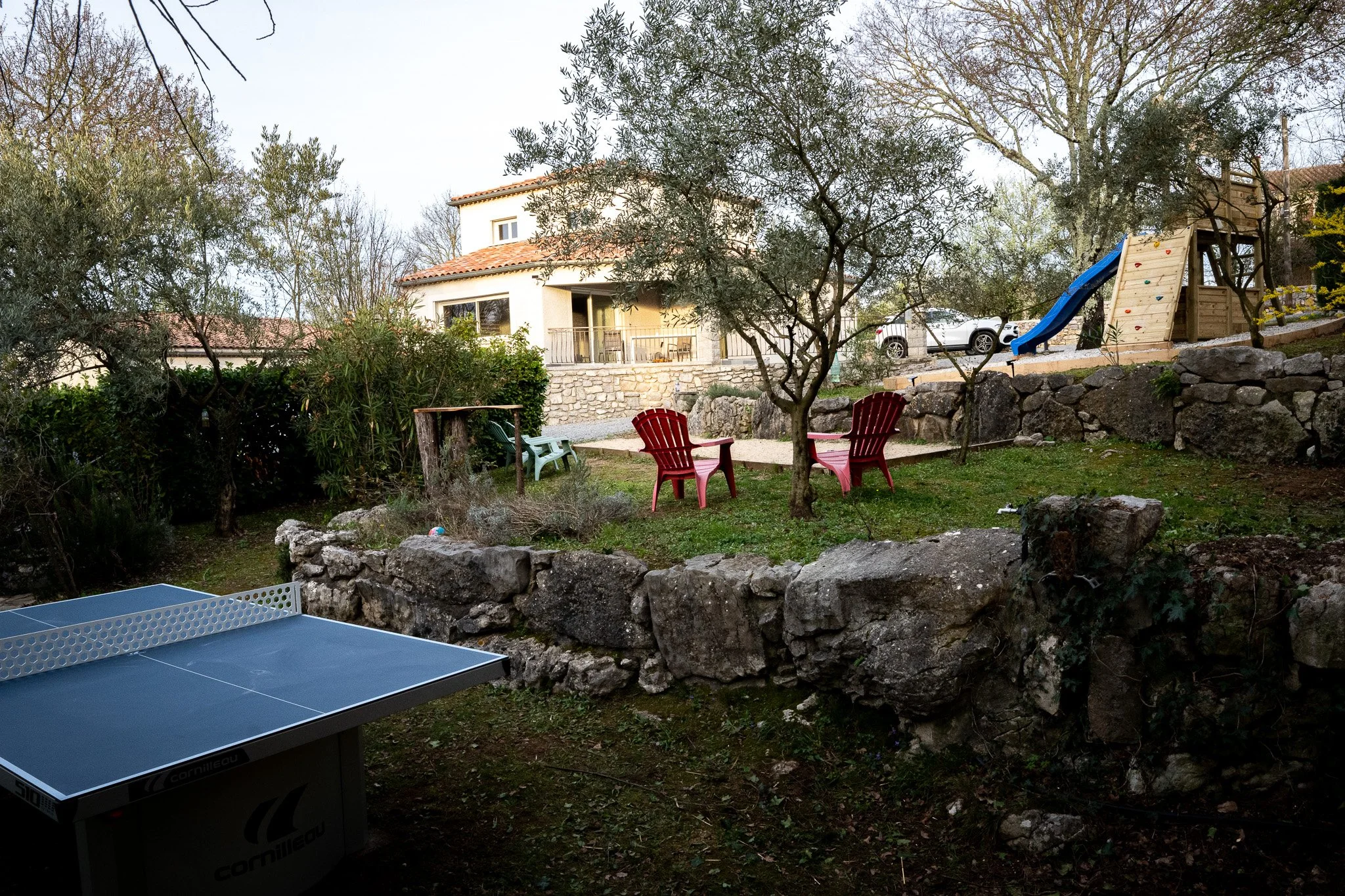 Jardin avec table de ping-pong, fauteuils rouges, une petite maison, un toboggan avec mur d'escalade, arbres et rochers.