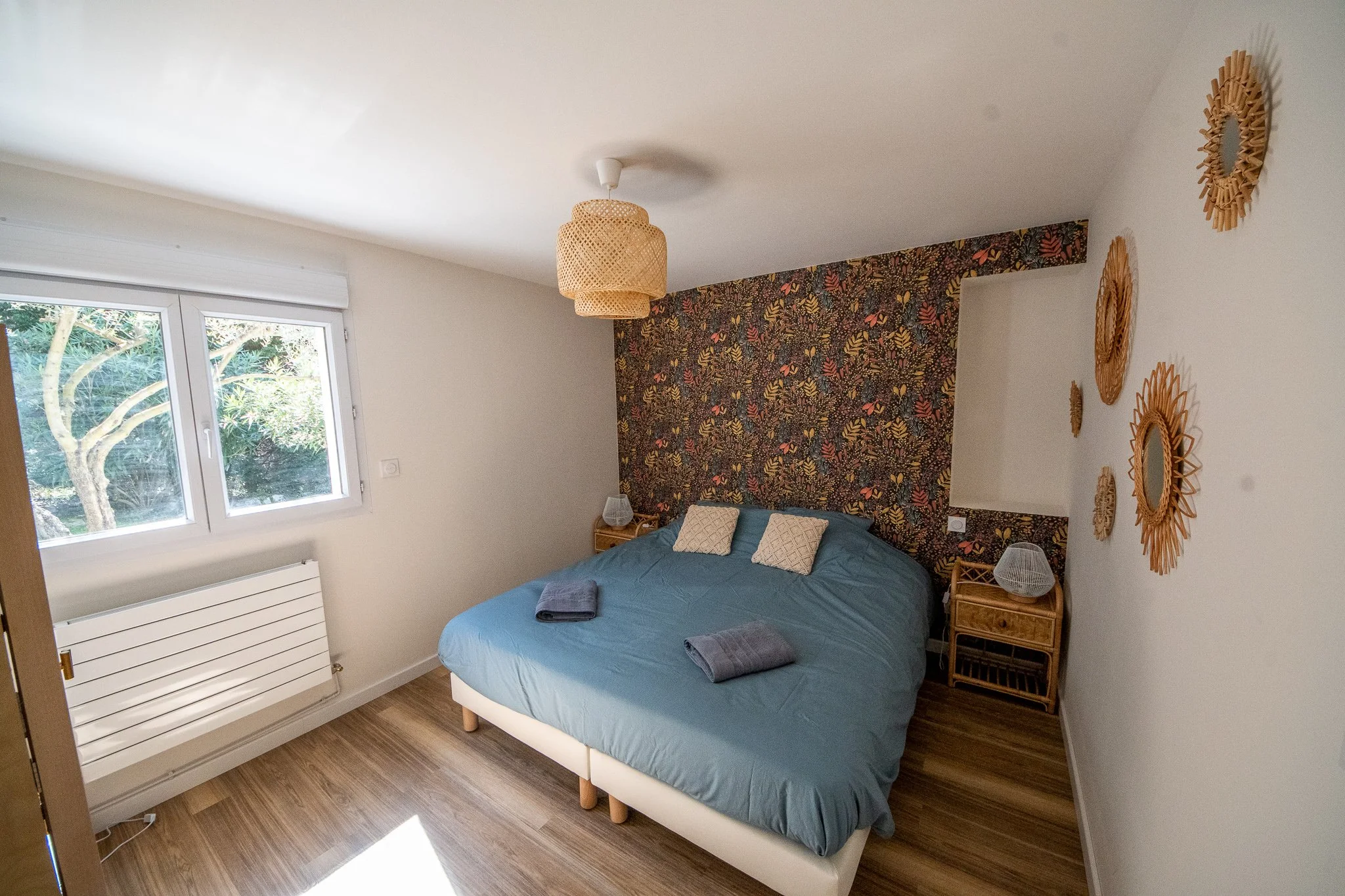 Chambre avec lit double, tapisserie floral derrière le lit, deux lampes de chevet, trois serviettes sur le lit, plusieurs miroirs en rotin sur le mur, une fenêtre avec vue sur la nature.