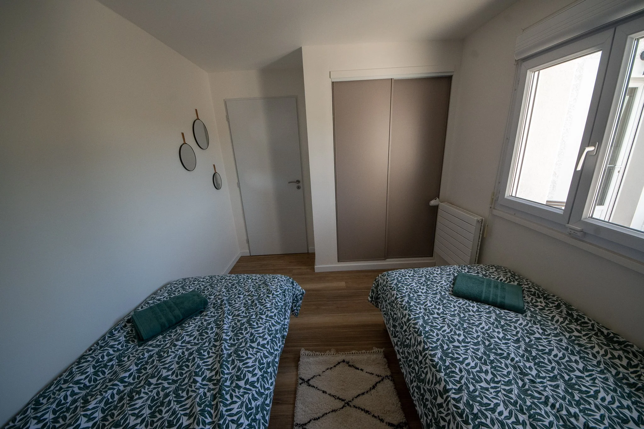 Chambre à deux lits avec draps à motifs végétaux, une fenêtre laissant entrer de la lumière naturelle, un placard à portes coulissantes, un radiateur, trois petits miroirs ronds sur le mur, et un tapis beige avec motifs noirs.