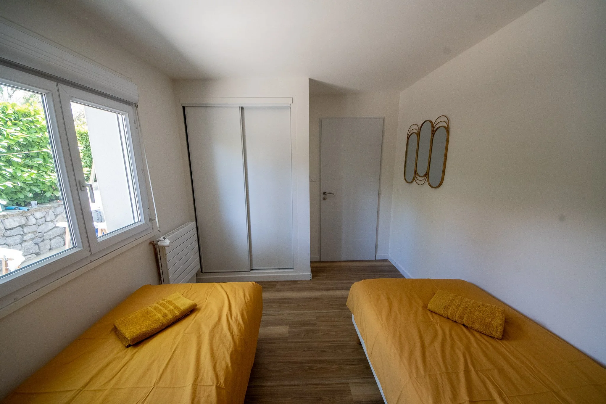 Chambre avec deux lits simples avec draps jaunes et serviettes jaunes, fenêtre avec vue sur jardin, placard blanc et décorations murales en rotin.